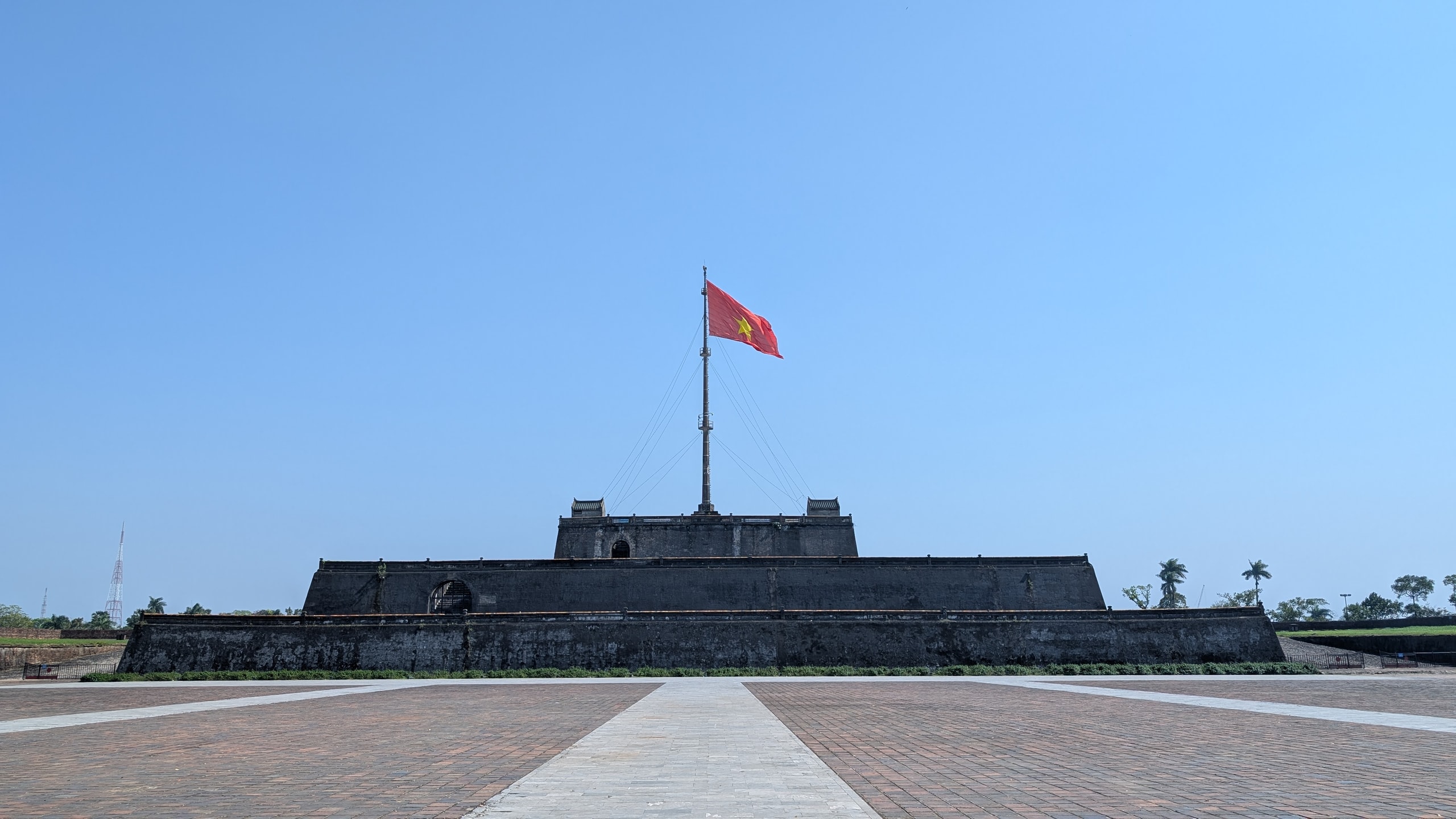 The Flag Tower of Hue, a three-tiered dark brick platform with a large red Vietnamese flag on a tall pole, seen across an empty paved square