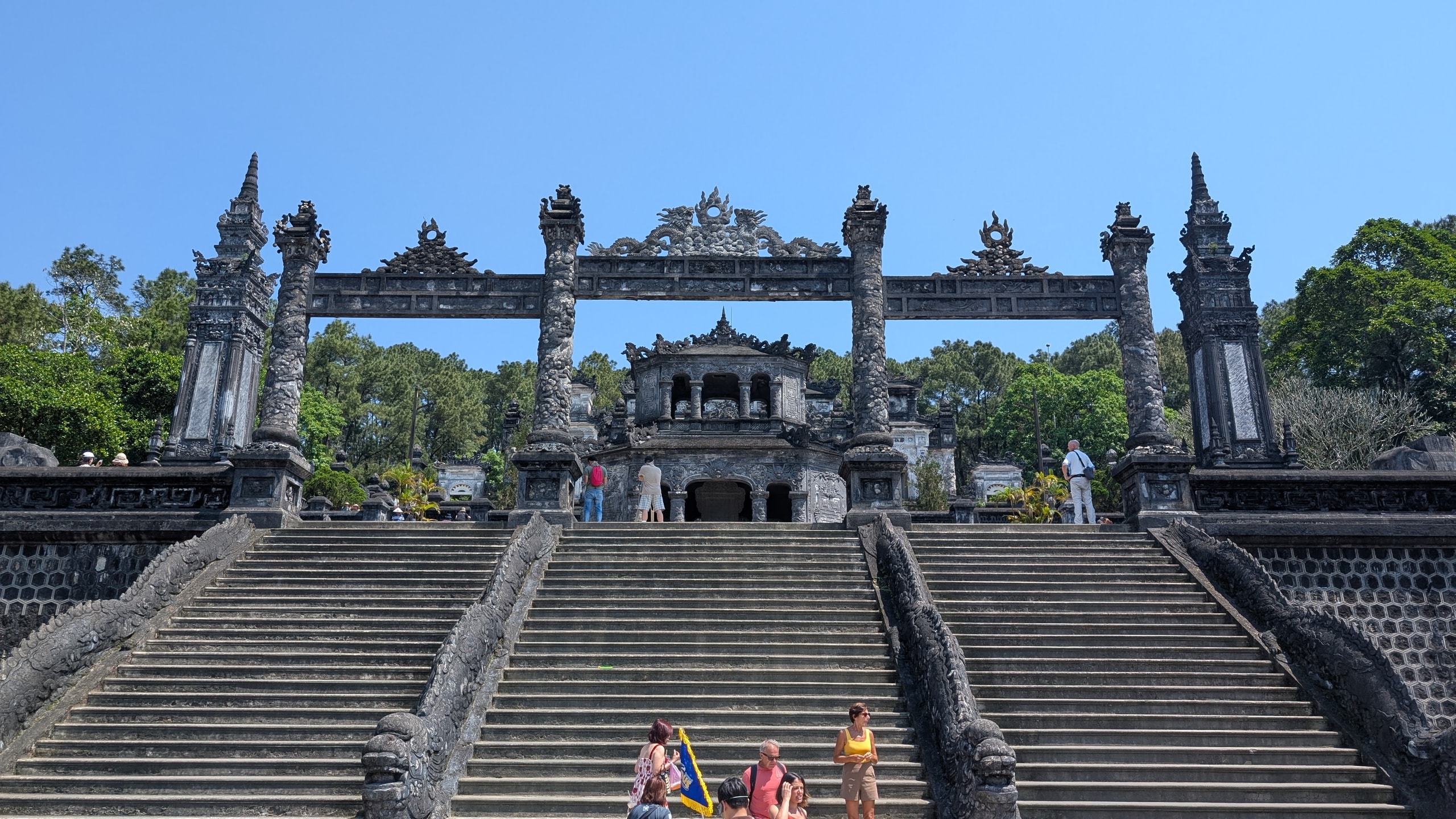Three flights of stone stairs with dragon-carved banisters leading up to a dark concrete gateway and pavilion at Khai Dinh's tomb, with a tour group at the bottom