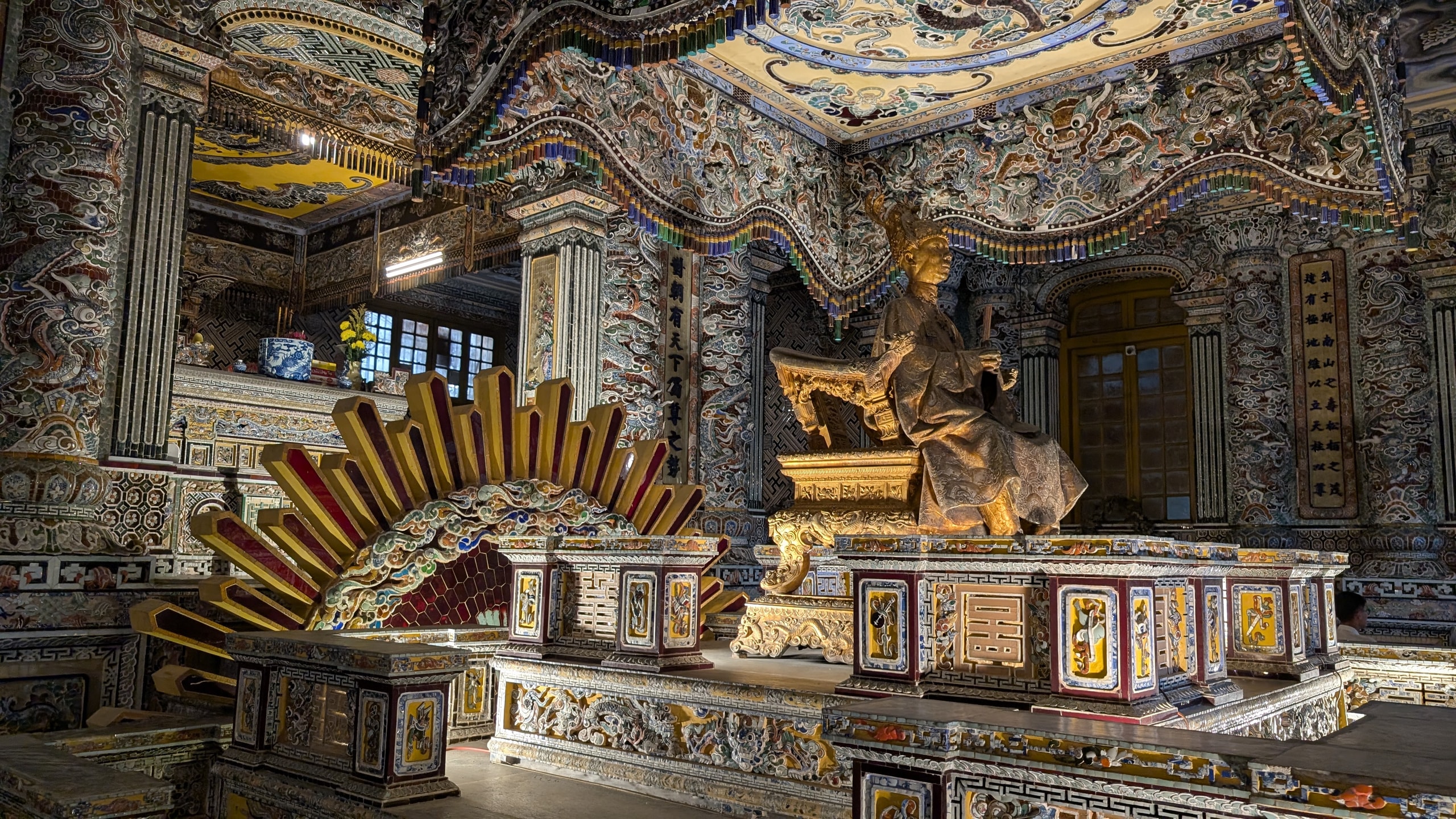 Inside Thien Dinh hall at Khai Dinh's tomb, a gilded seated statue of the emperor under a canopy, surrounded by walls and columns covered in porcelain and glass mosaic