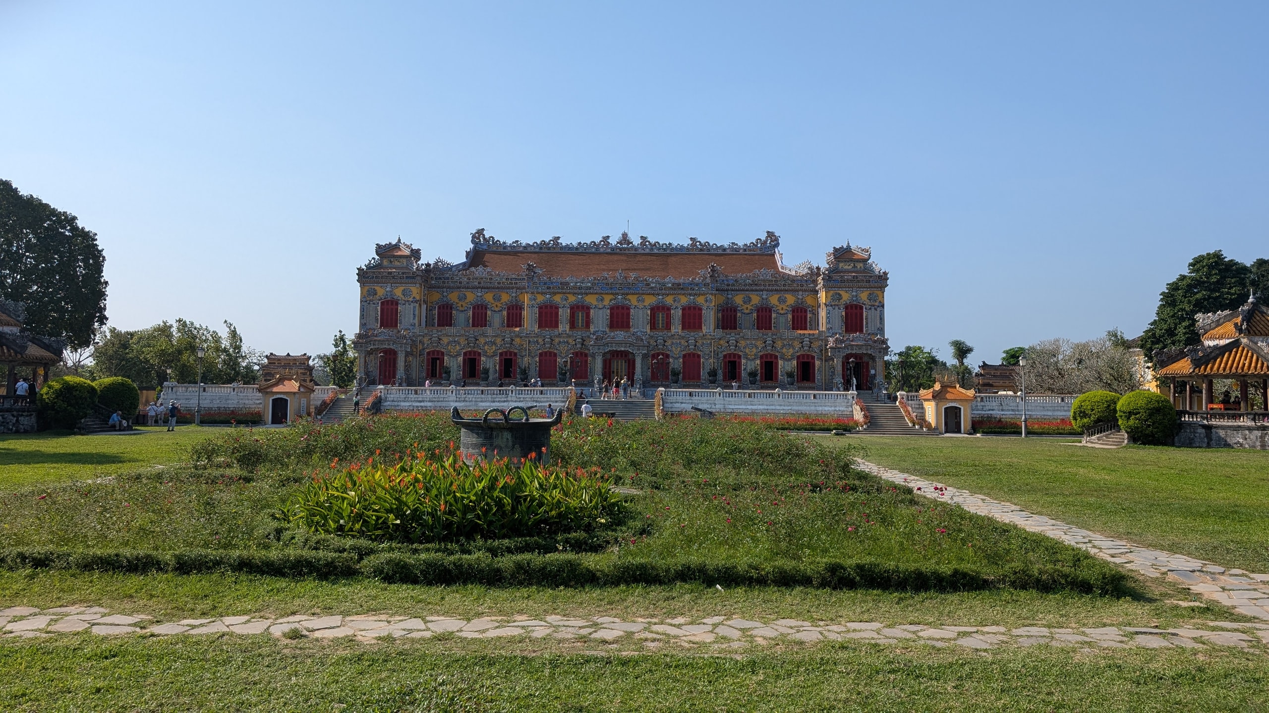 Kien Trung Palace, a two-storey yellow European-style palace with rows of red shutters and ornate rooftop decoration, seen across a lawn and flower beds in Hue's Imperial City