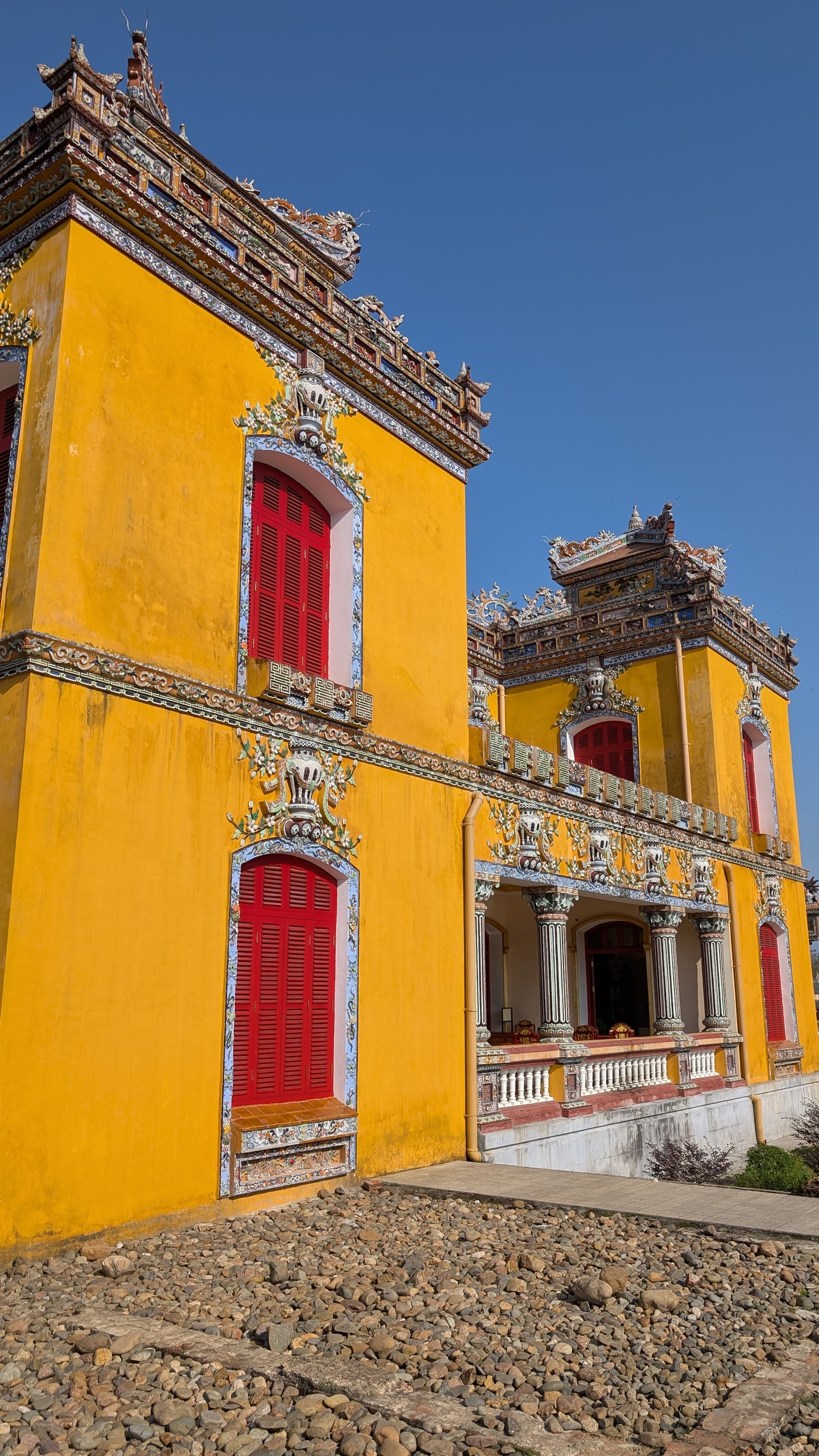 The side of Kien Trung Palace with deep yellow walls, red wooden shutters framed in ceramic trim, and a pillared veranda under an ornate roof