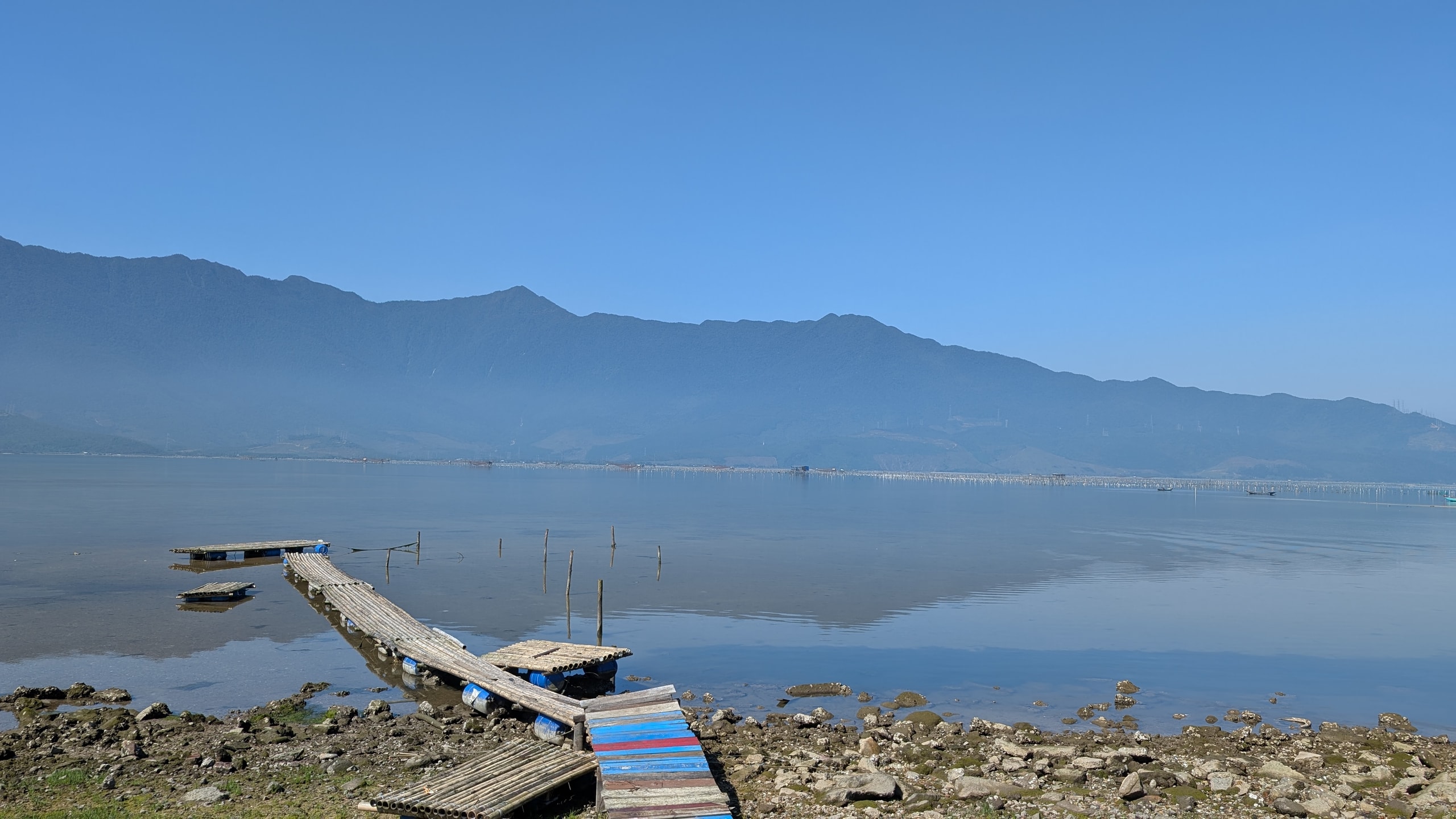 Flat water of Lap An lagoon at Lang Co with a painted wooden walkway on blue floats in the foreground and a long mountain ridge behind