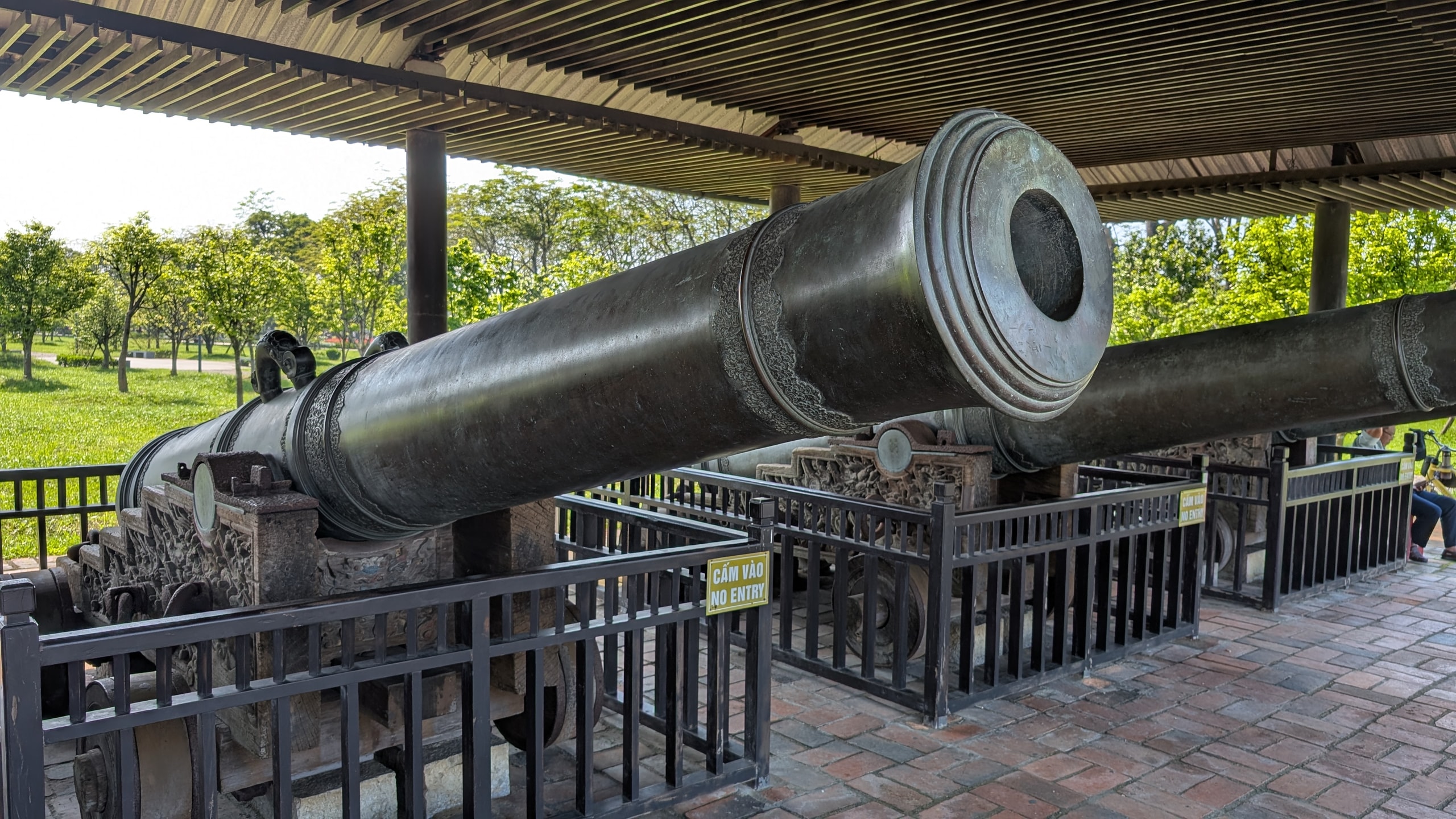 Two long bronze cannons with carved bands on stone carriages, fenced off under a covered pavilion with green lawns and trees beyond