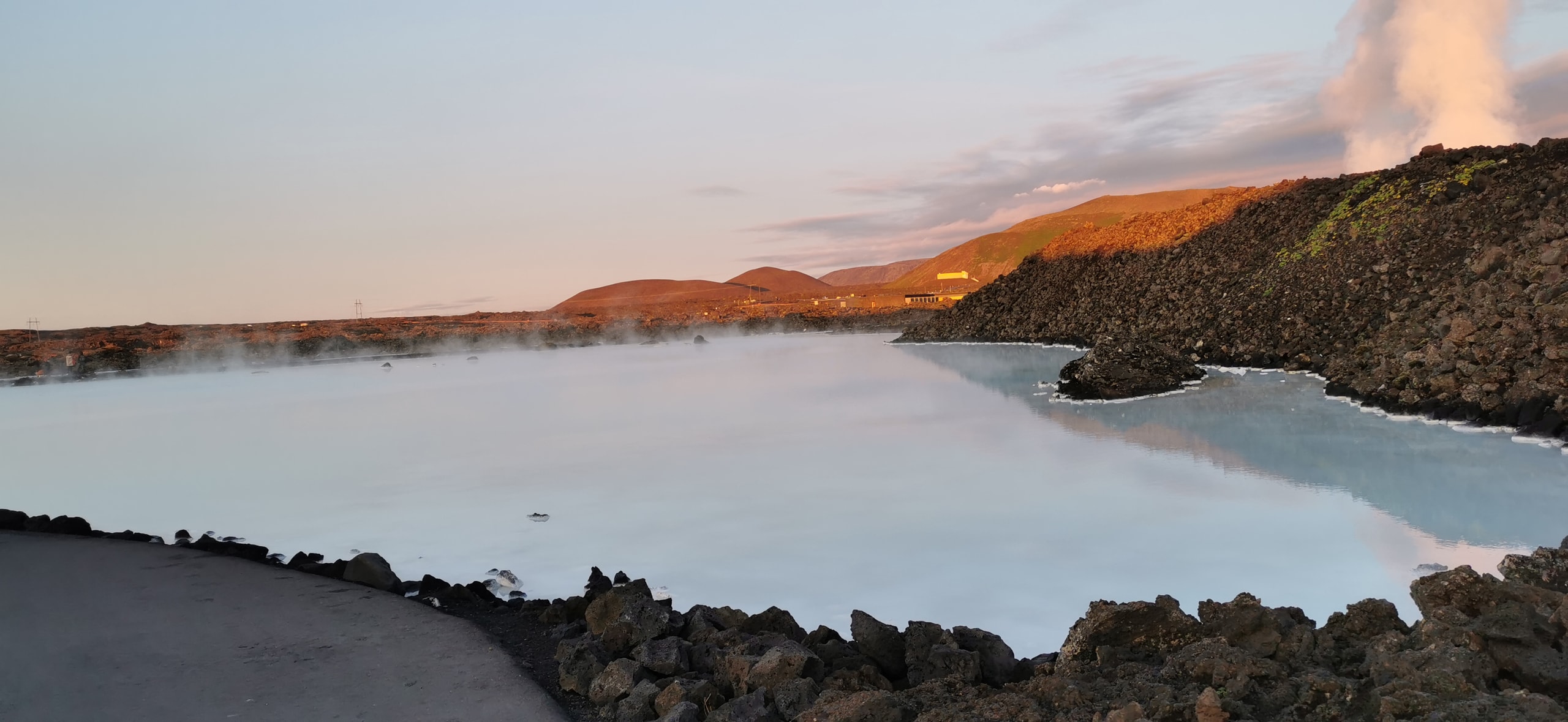 Milky blue Blue Lagoon water among black lava rocks with steam rising and pink evening light on distant hills