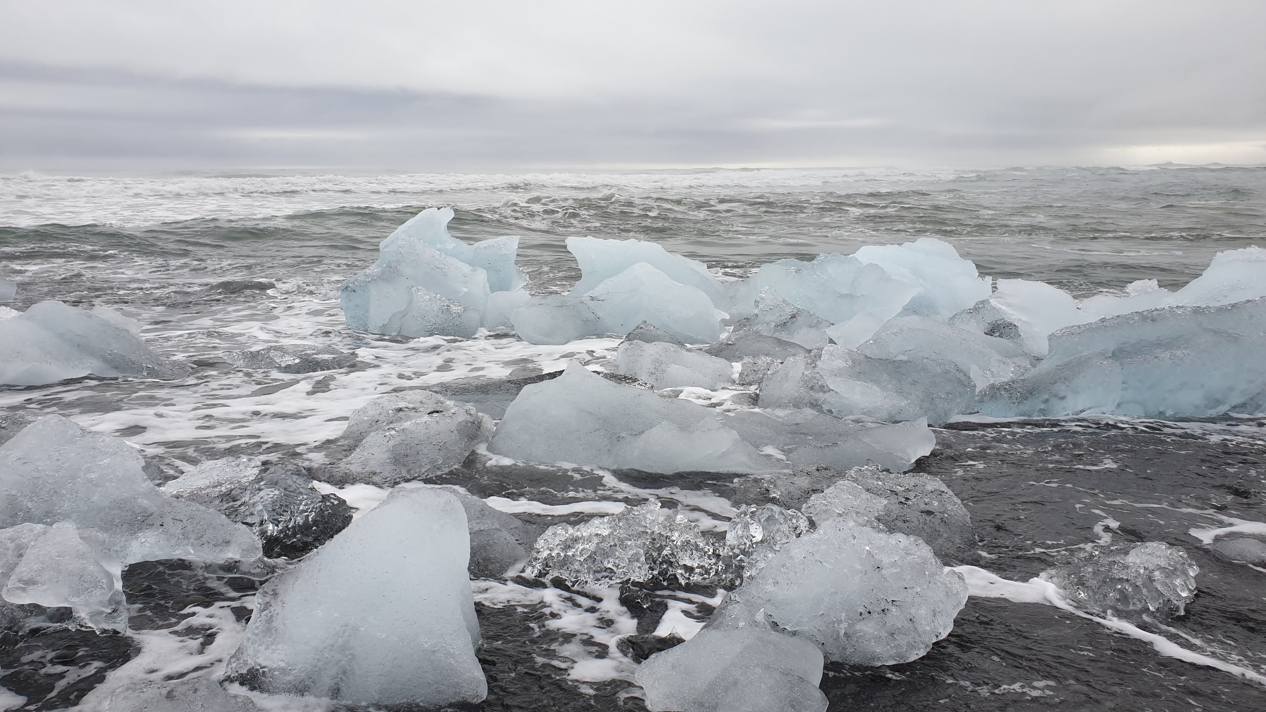 Chunks of white and blue glacial ice scattered on wet black sand at Diamond Beach with foamy waves and a grey ocean horizon