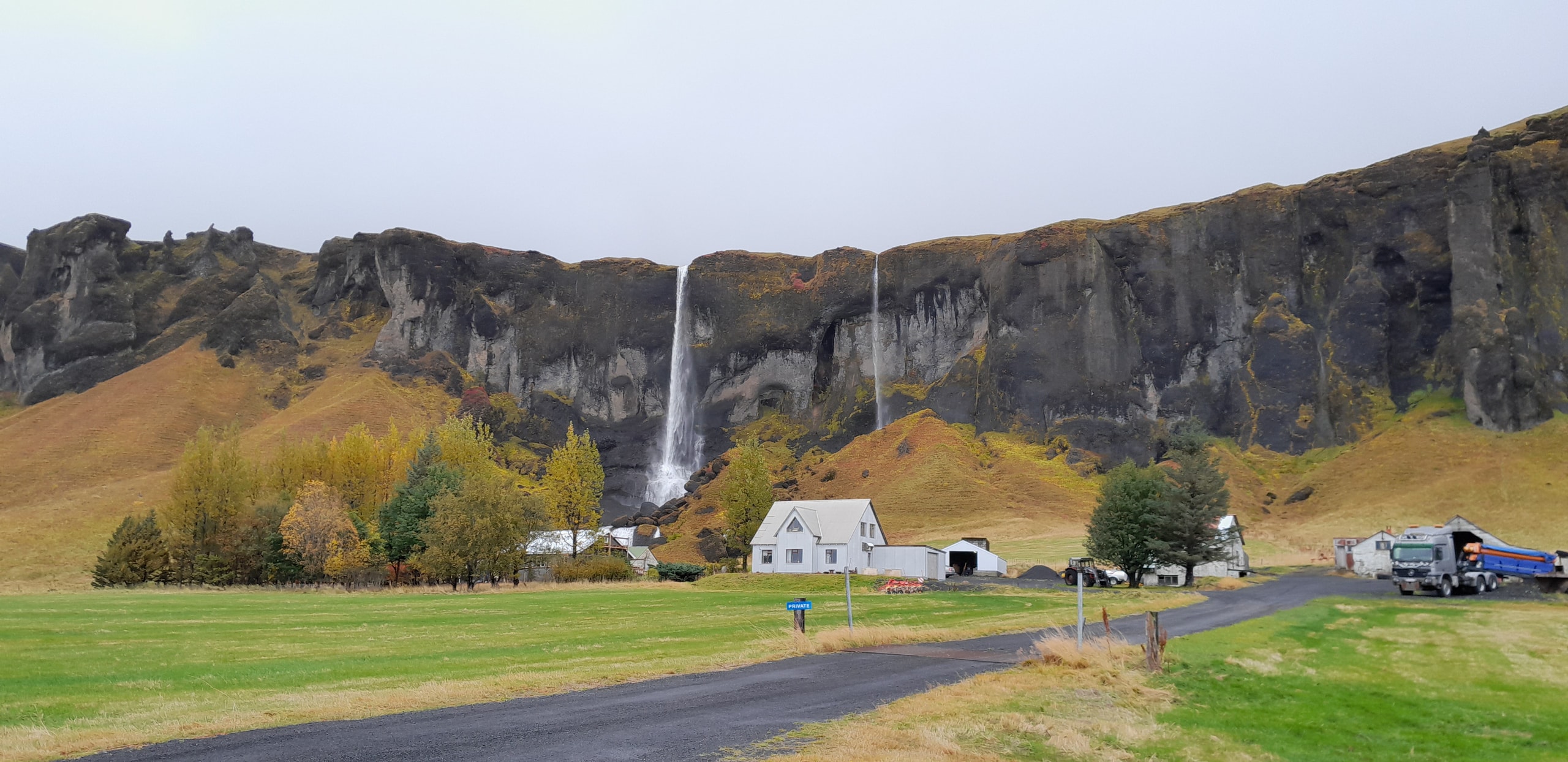 White farmhouse and outbuildings beneath a dark cliff with two waterfalls, green fields, and a blue PRIVATE sign by the driveway