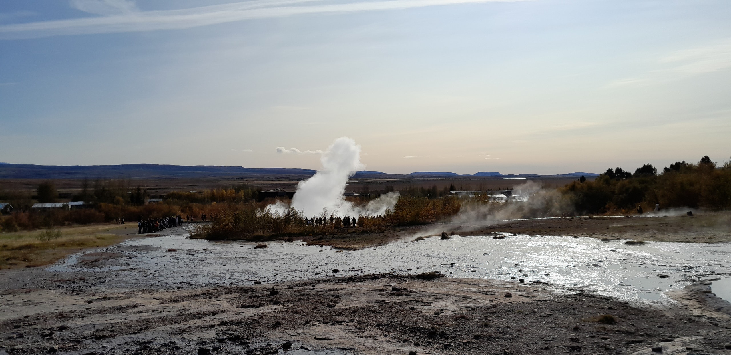 Strokkur geyser erupting in a white plume of steam while a crowd of visitors stands in a circle on the wet geothermal ground
