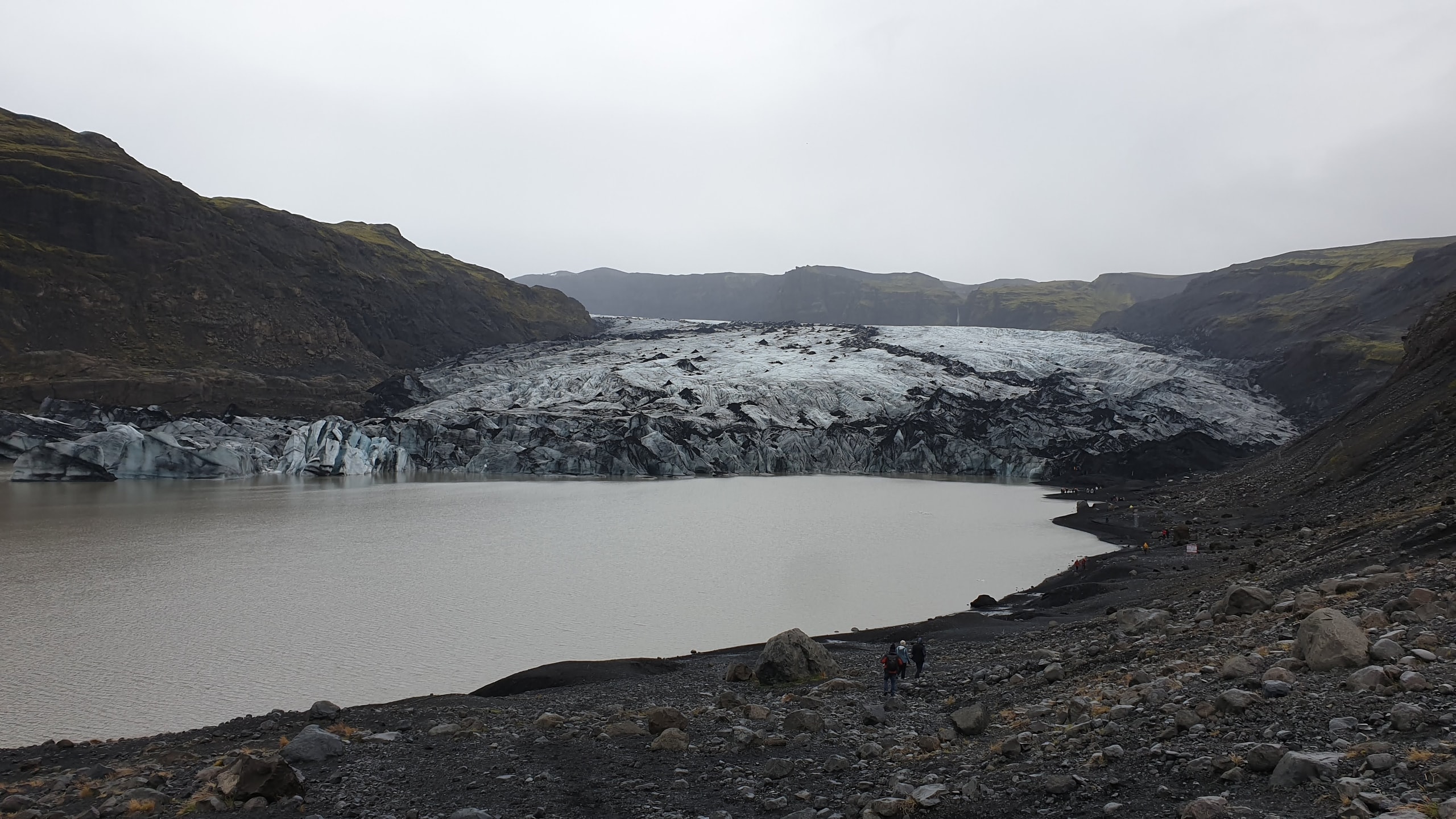 Sólheimajökull glacier with black volcanic ash streaks above a brown meltwater lagoon and hikers on the rocky shoreline