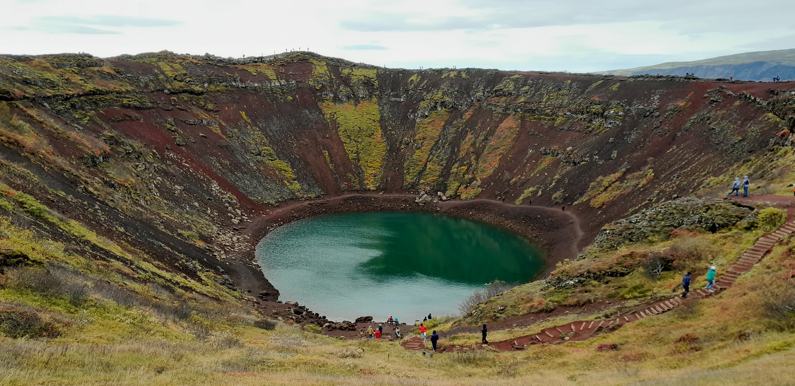 Kerið volcanic crater with rust-red slopes, green moss, and a teal lake at the bottom with visitors on the rim path
