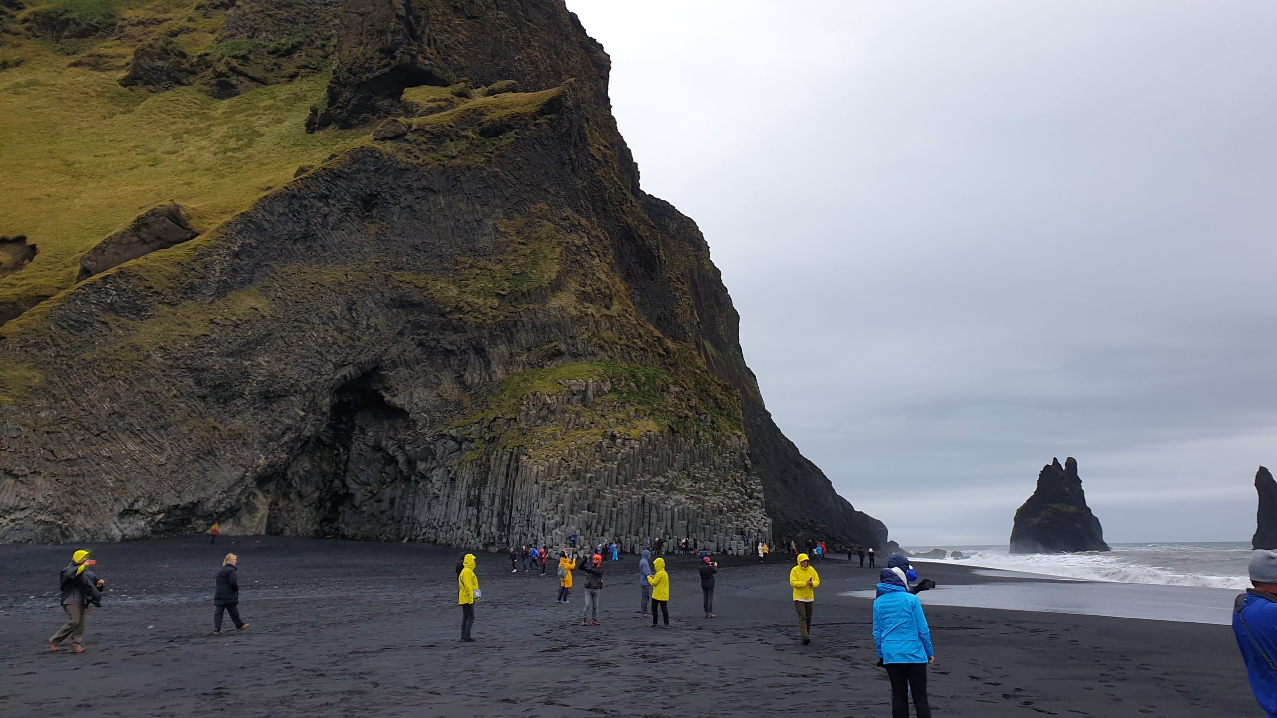 Tourists in bright rain jackets on Reynisfjara black sand beach beside hexagonal basalt columns and sea stacks under grey sky