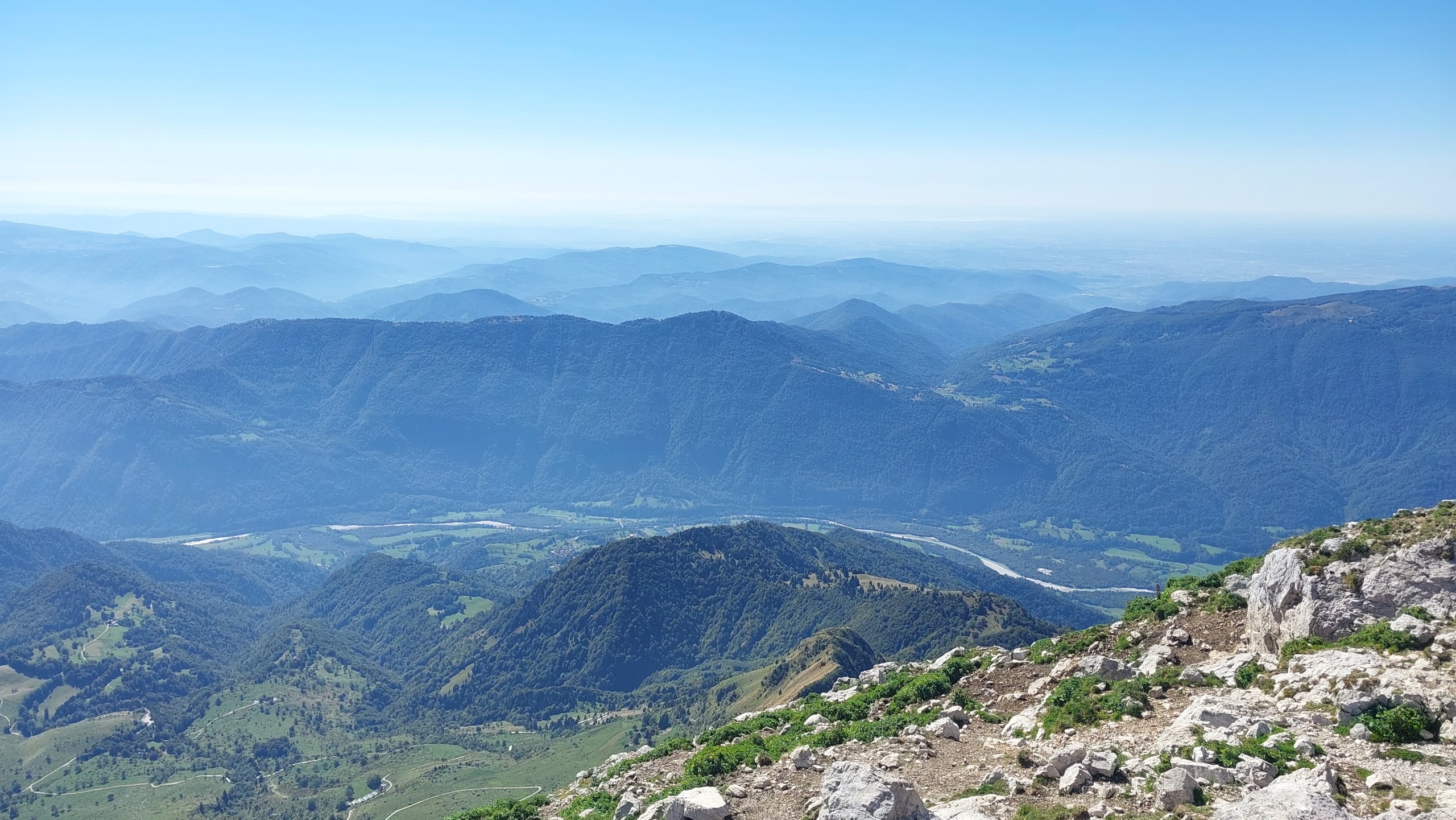View south from the rocky upper slopes of Krn over green hills, the braided Soča river, the Kolovrat ridge and a hazy plain beyond