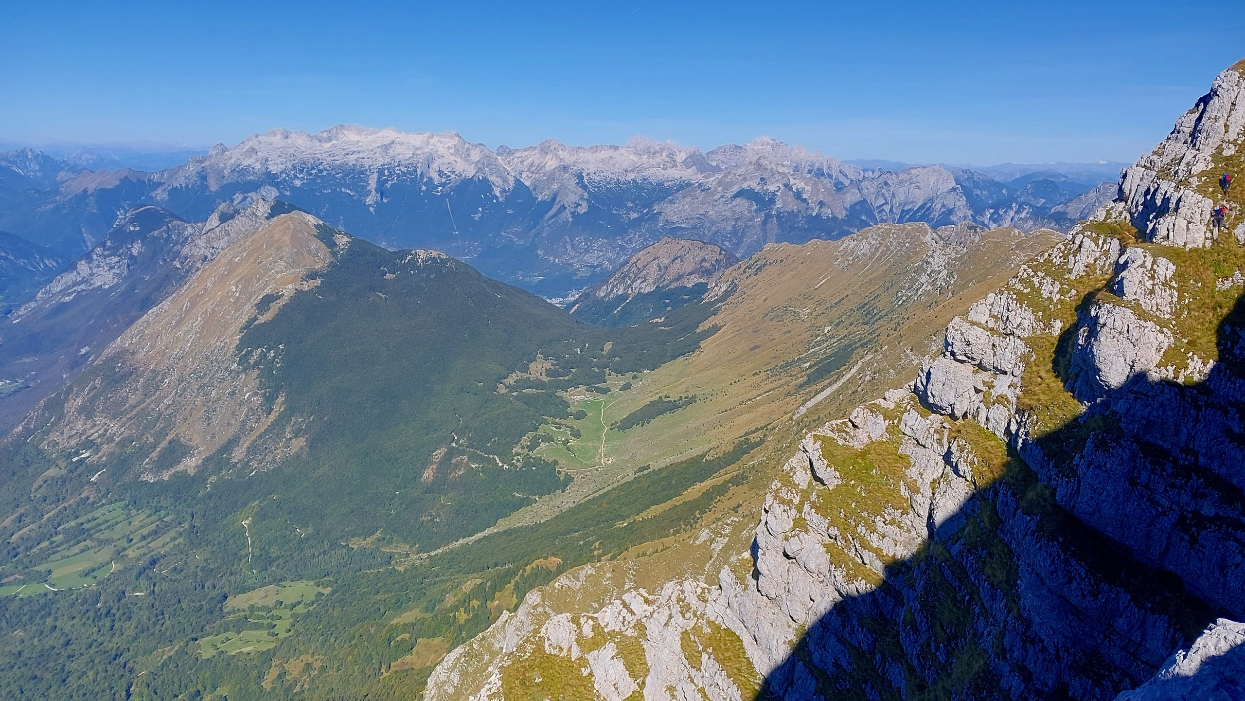 Grassy limestone ridge of Krn falling away to the west, with two hikers on the crags and the pale Kanin range above the Bovec basin