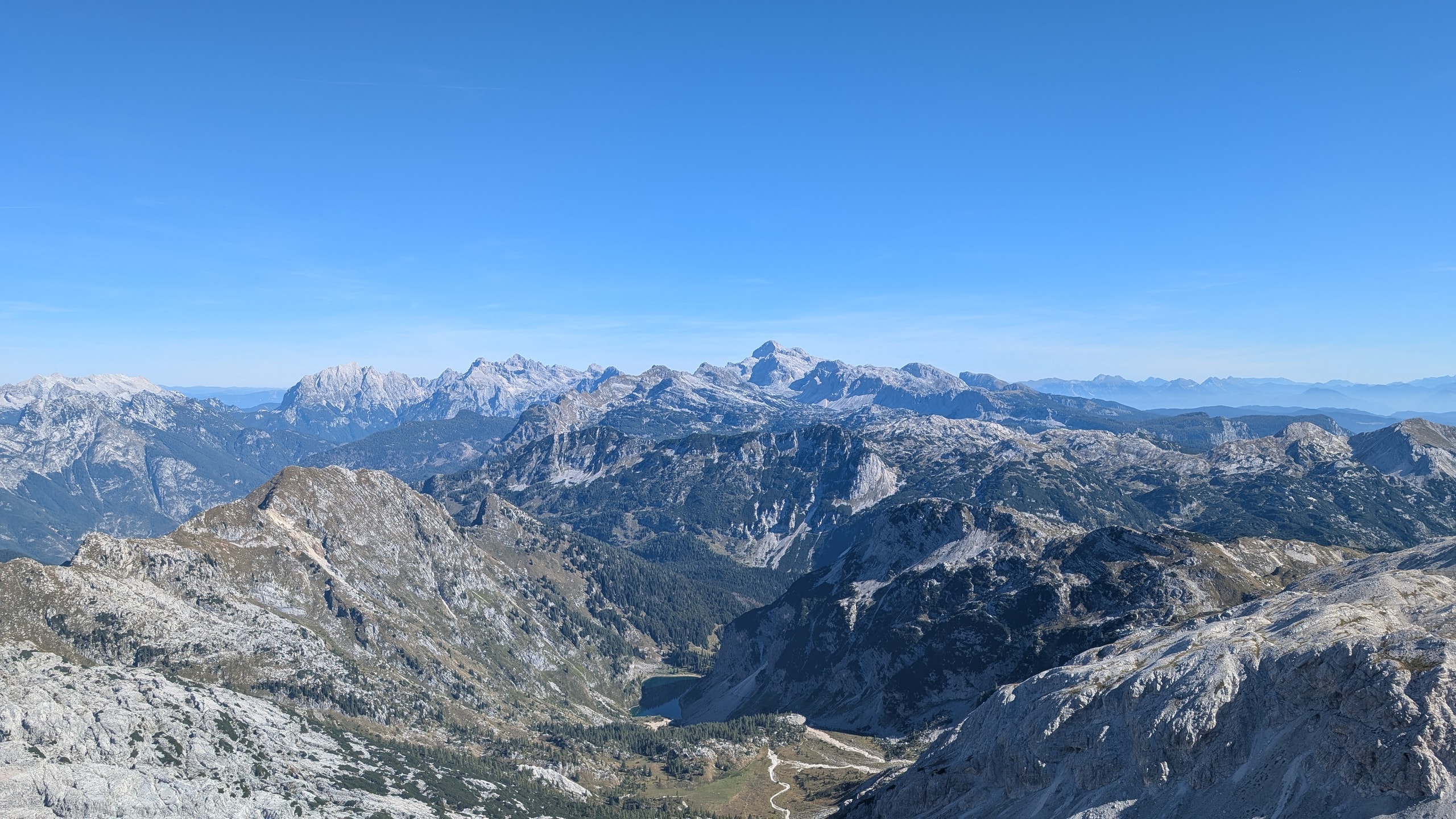 View from the summit of Krn over grey limestone ridges to Triglav on the skyline, with dark Krn Lake in a forested bowl far below