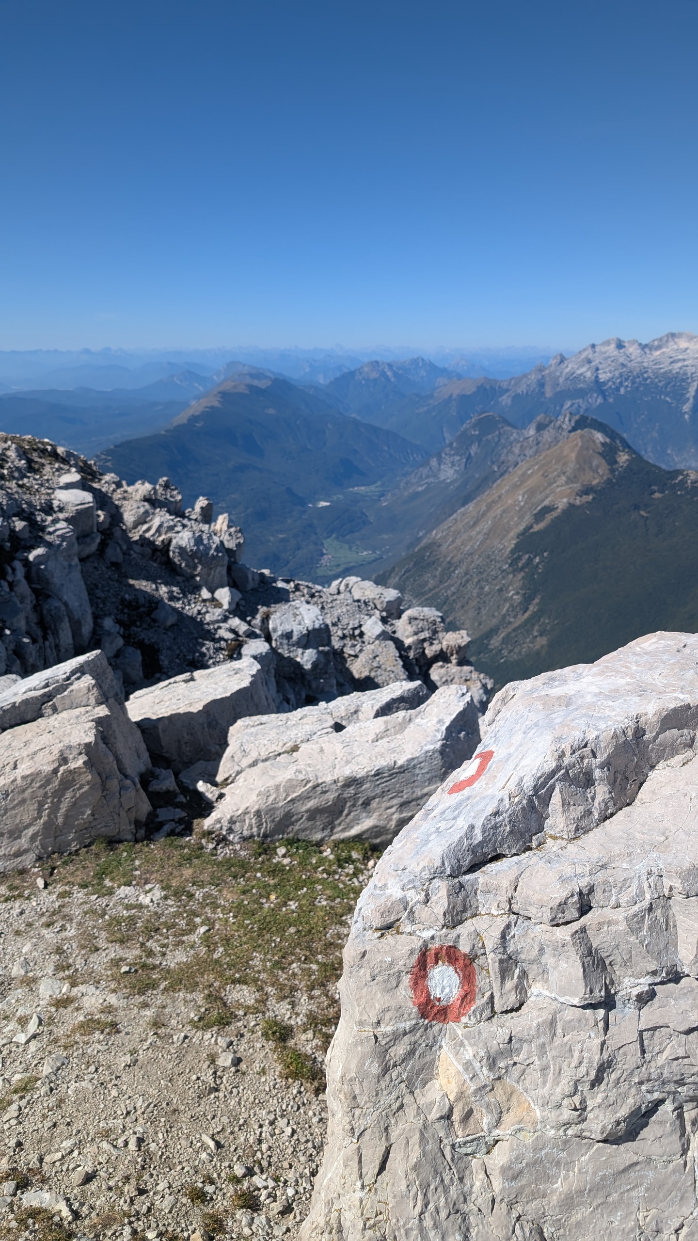 Red and white circular trail mark painted on a limestone boulder near the summit of Krn, with the Soča valley and mountains beyond
