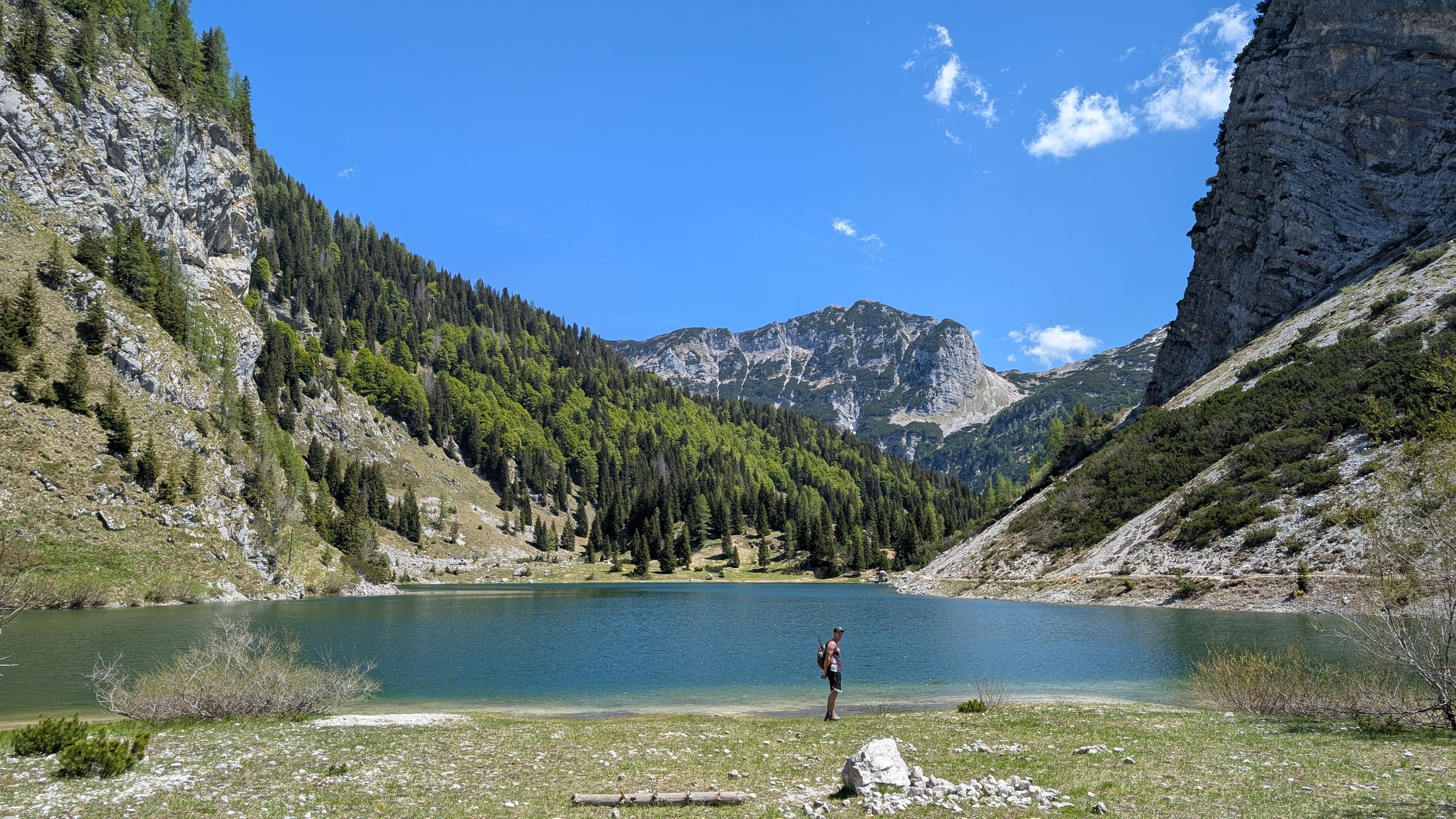 Hiker with a backpack standing on the grassy shore of turquoise Krn Lake with limestone peaks beyond