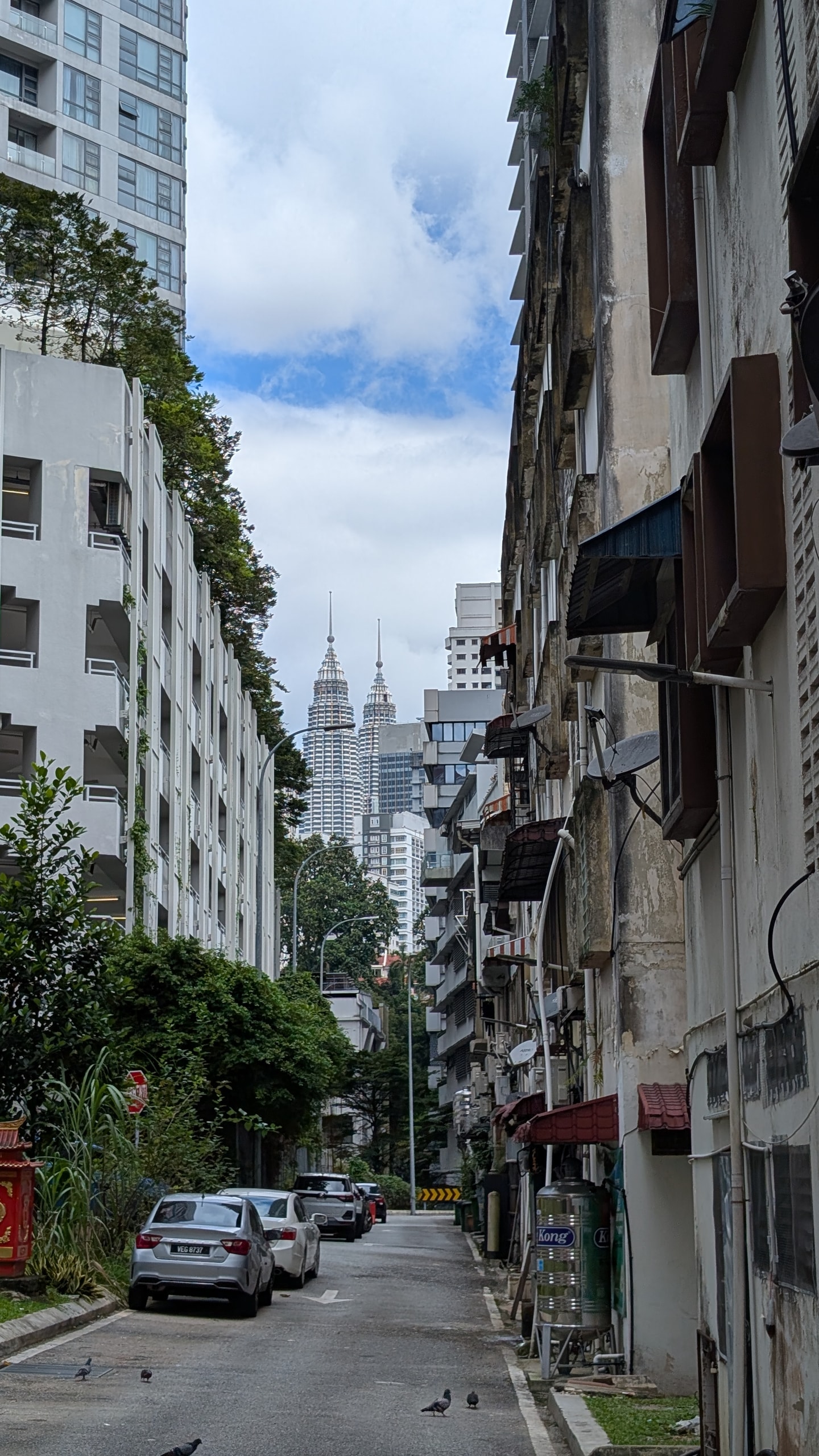 A narrow back lane with weathered shophouse walls, a steel water tank, parked cars and pigeons, the Petronas Twin Towers visible at the far end
