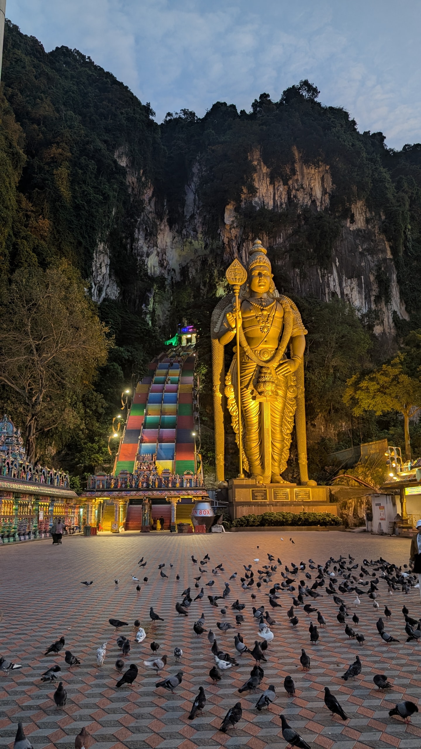 The gold Lord Murugan statue and the rainbow-painted stairs at Batu Caves before sunrise, with pigeons on the empty tiled plaza below a limestone cliff