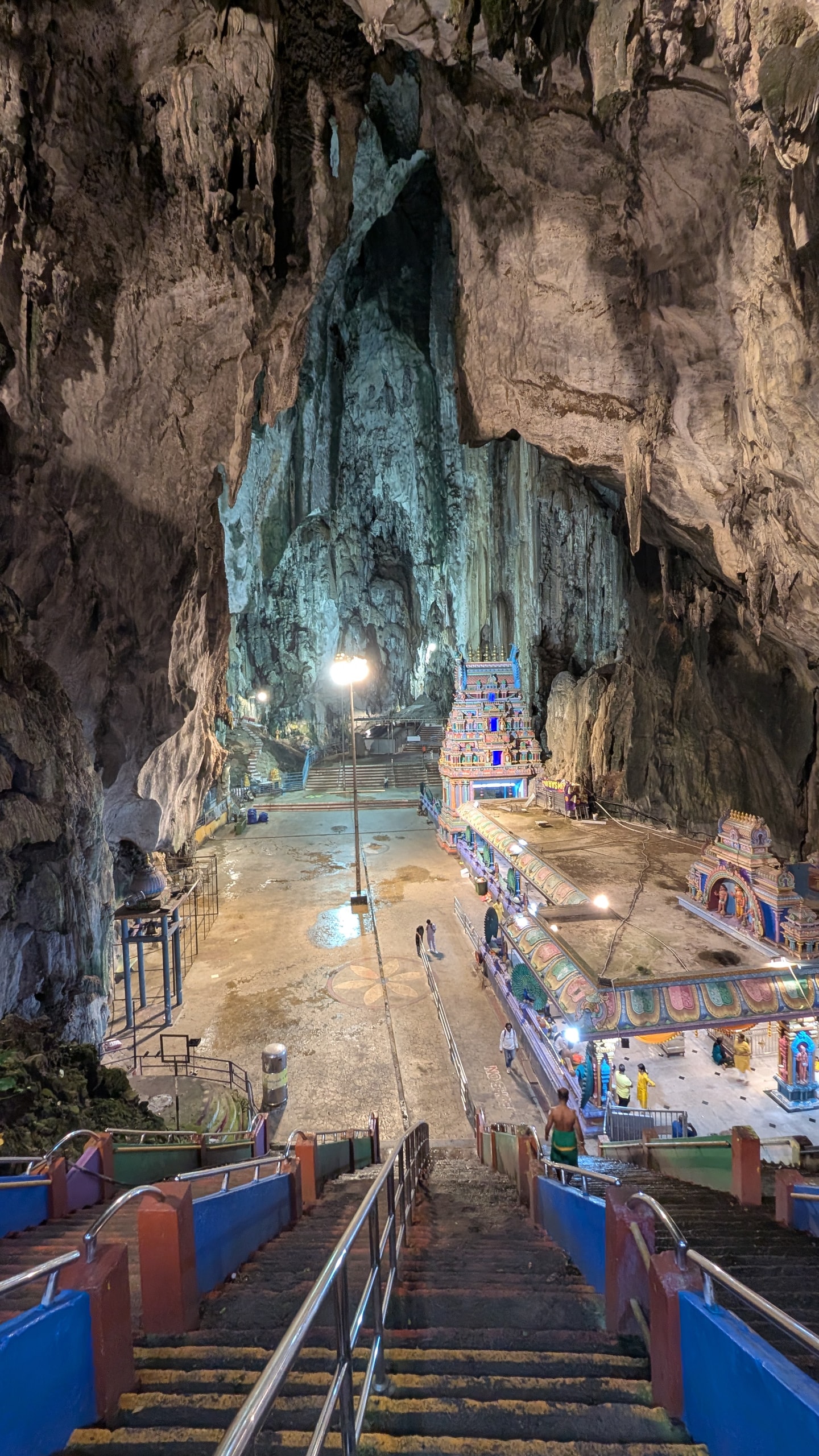 View down the stairs into the Temple Cave at Batu Caves, with a lit painted shrine, a lamp post and a wet stone floor under a huge limestone ceiling