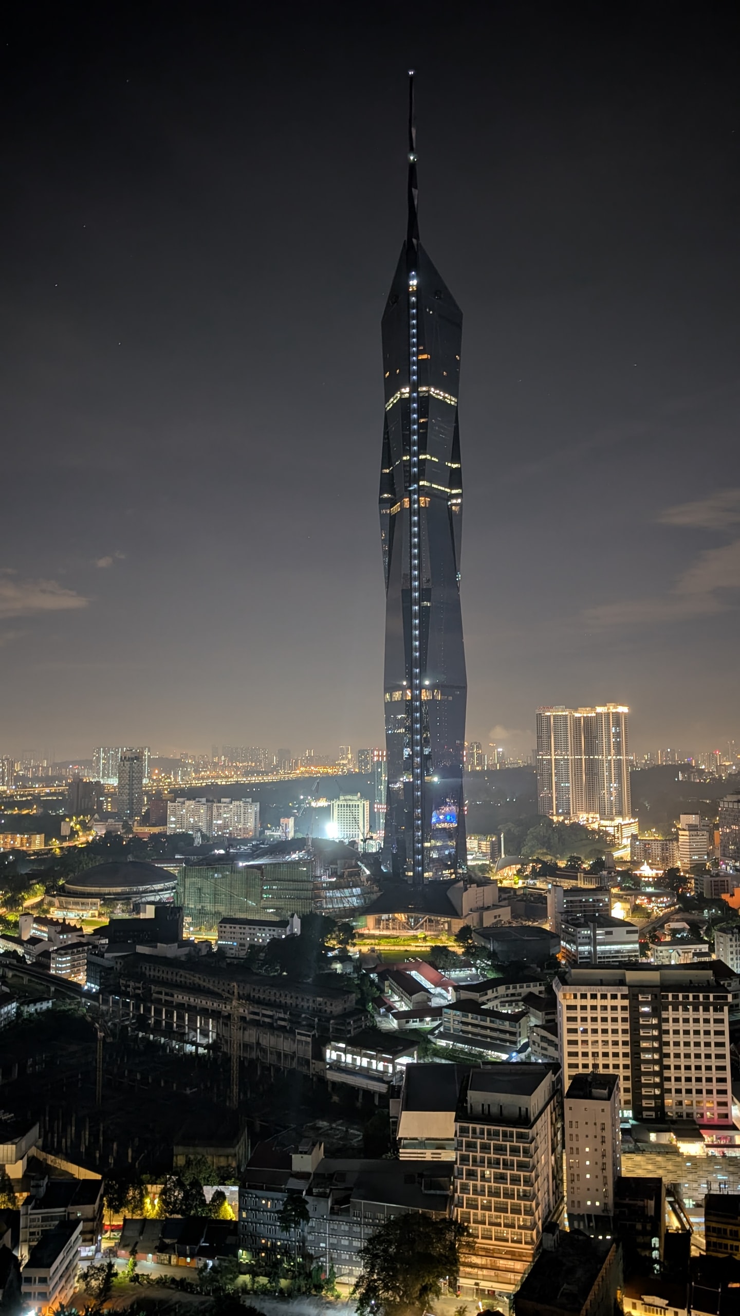 Merdeka 118 at night, a tall faceted glass tower with a line of lights running up to its spire above the lit low-rise streets of central Kuala Lumpur