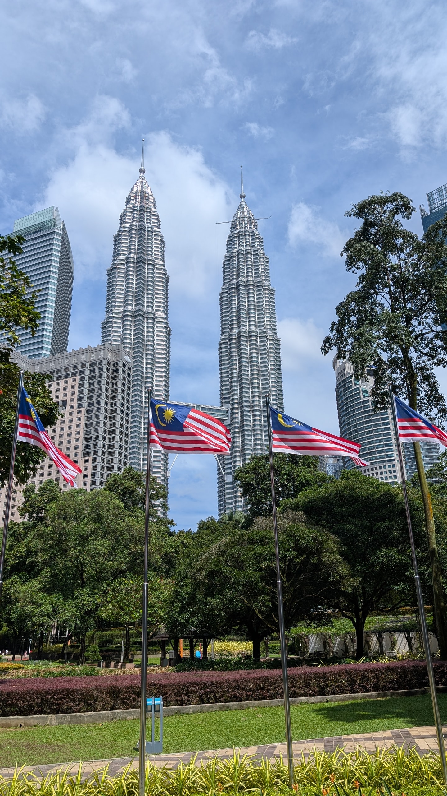 The Petronas Twin Towers rising behind a row of Malaysian flags and green trees in KLCC Park under a partly cloudy sky
