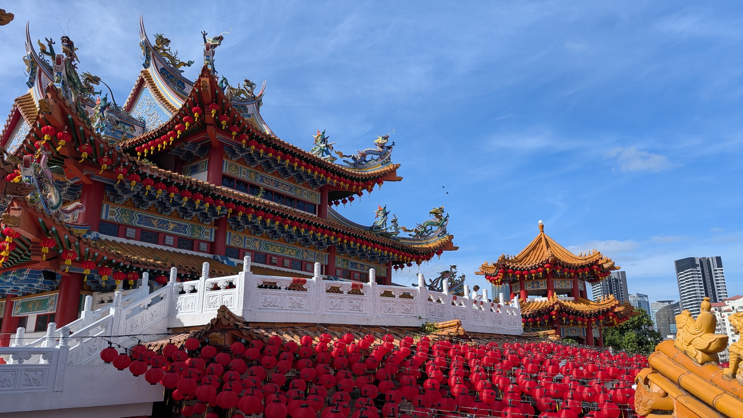 Red and gold roofs of Thean Hou Temple with dragon figures on the ridges above rows of red lanterns, a smaller pagoda and city towers to the right under a blue sky