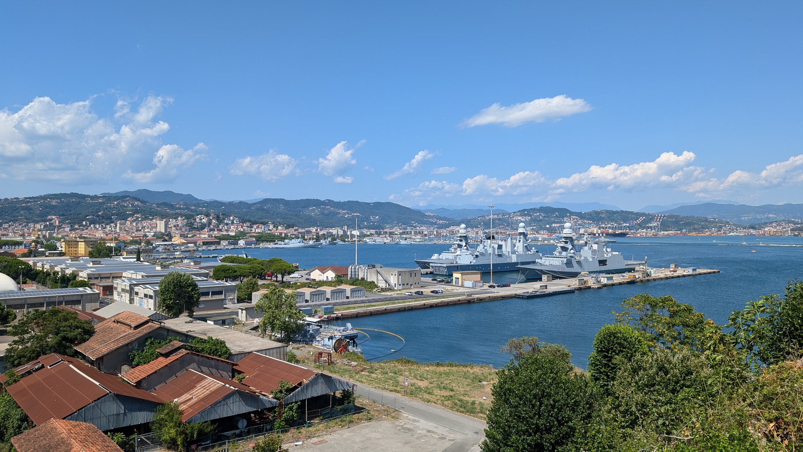 Grey navy frigates moored at a long pier in La Spezia harbour, with the city, port cranes, and green hills behind under a blue sky