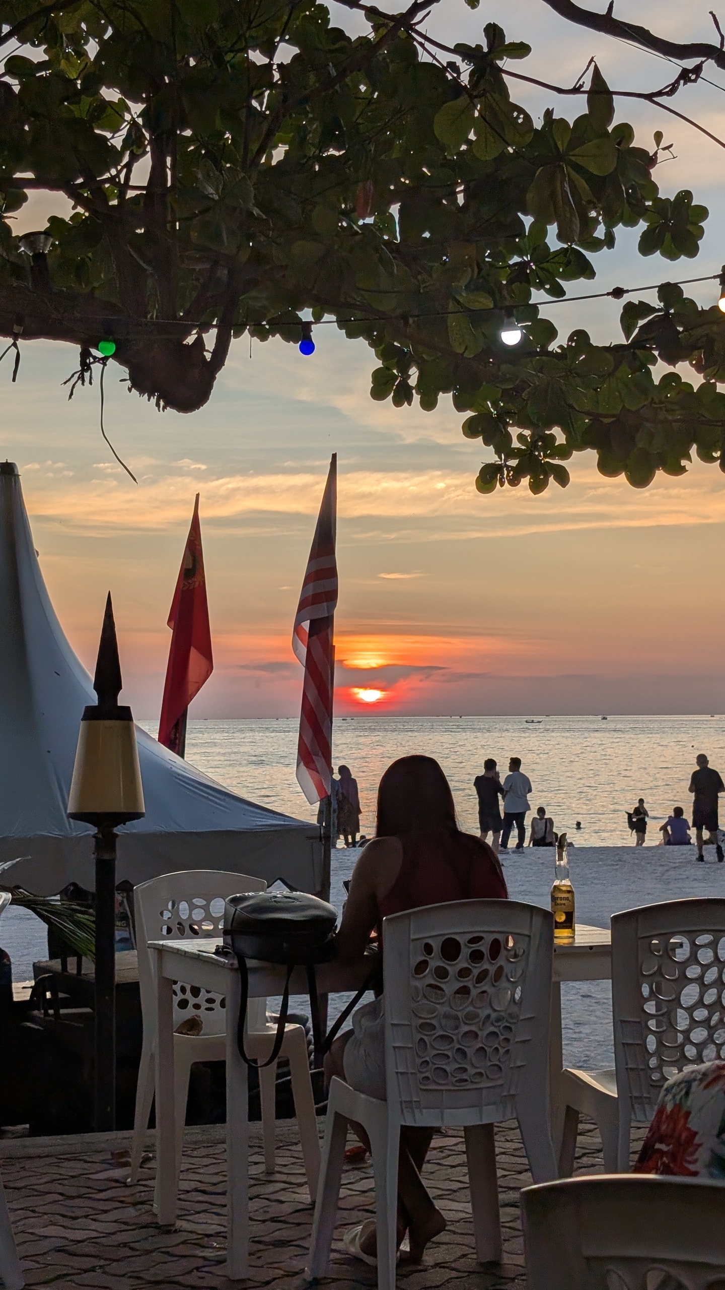 A table of white plastic chairs under string lights and a Malaysian flag, with a bottle of beer and the sun setting over the sea beyond