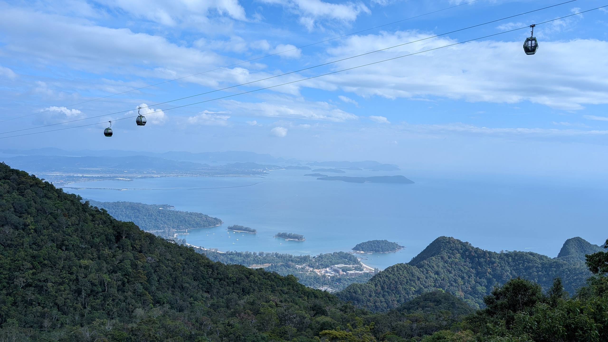 View from the Langkawi cable car over forested ridges to a pale blue bay dotted with small islands, two gondolas hanging from the cable