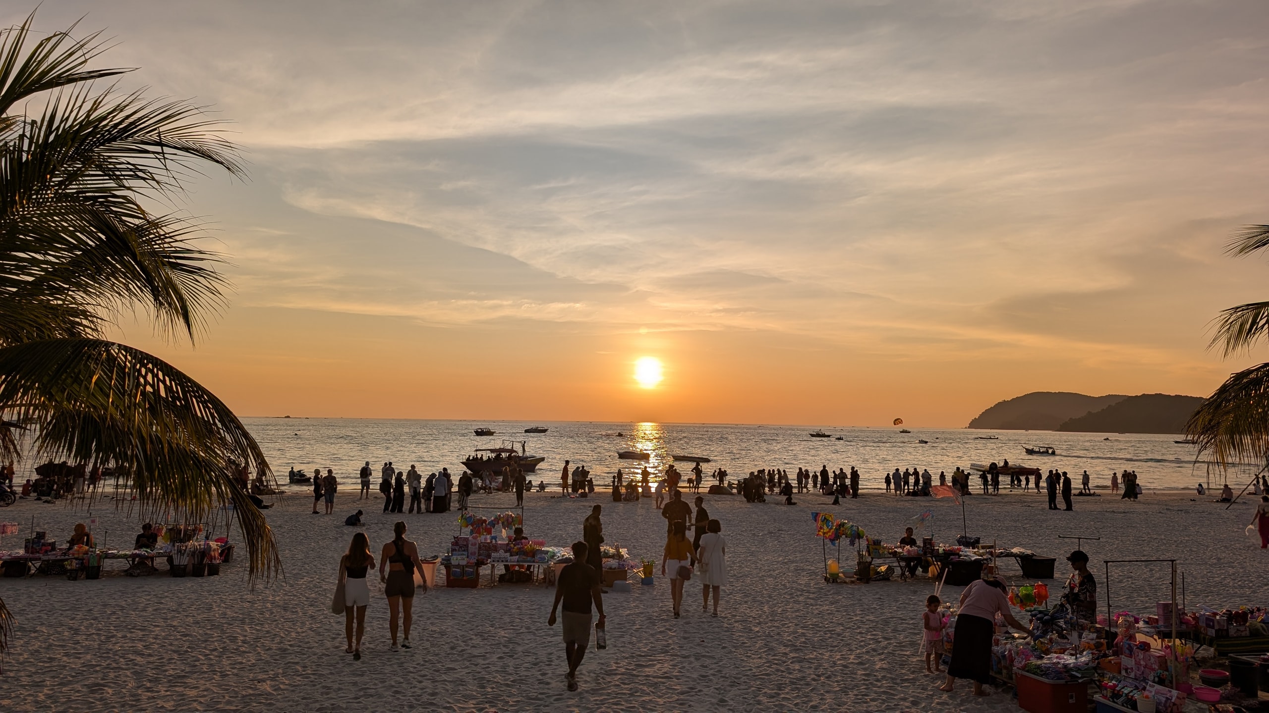 Crowds and toy stalls silhouetted on Pantai Cenang beach as the sun sets over the Andaman Sea with boats offshore