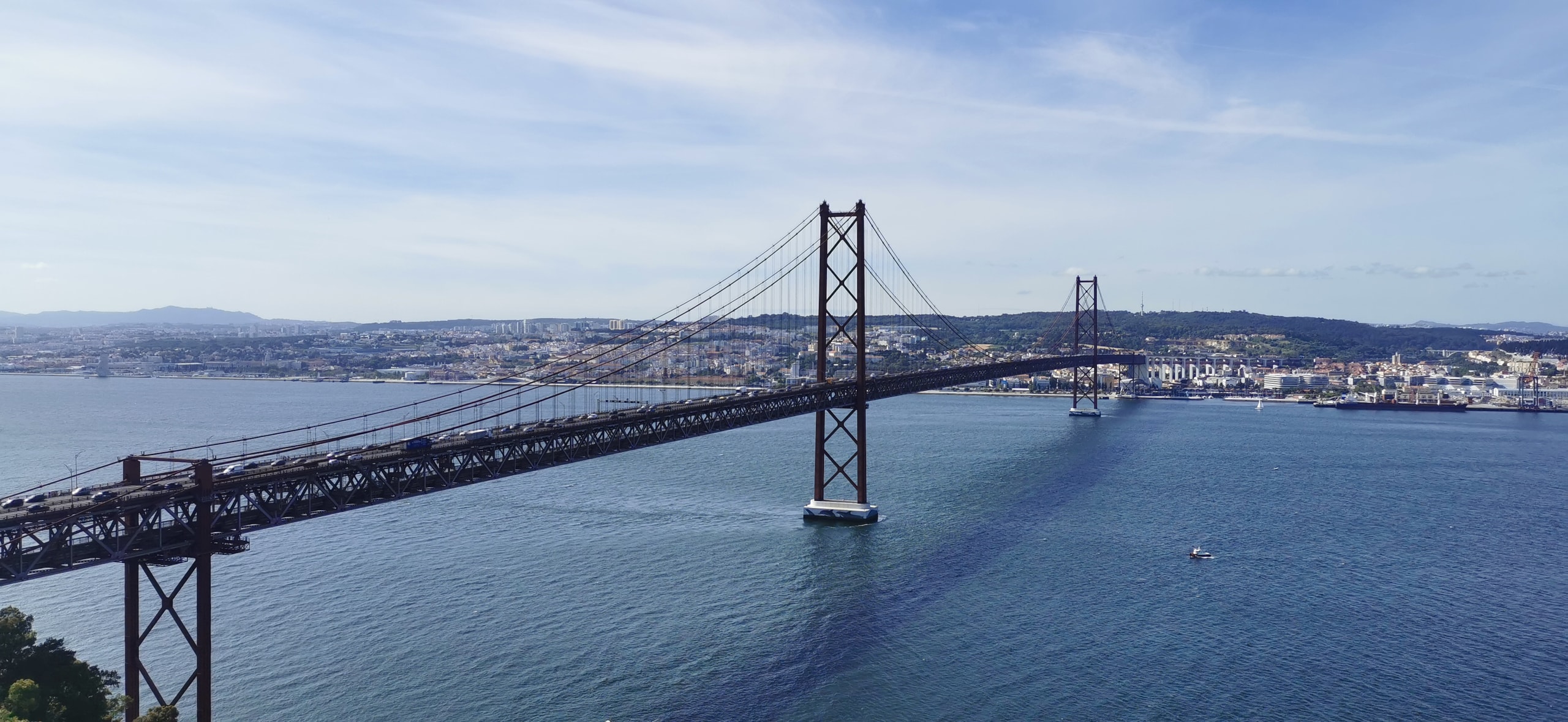 The rust-red 25 de Abril Bridge spanning the Tagus, with traffic on the deck, a small boat below, and Lisbon’s hills on the far bank