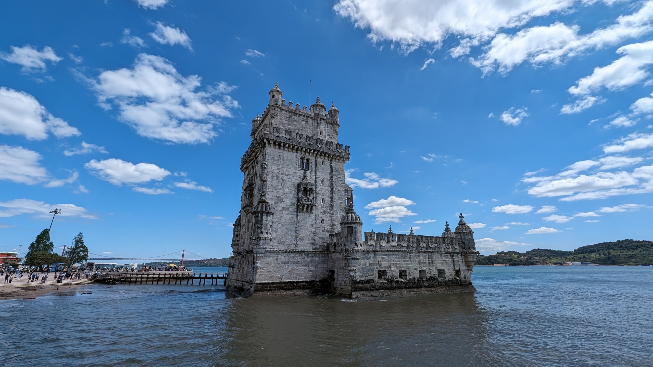 Belém Tower standing in the Tagus on a sunny day, with its wooden access bridge, onion-domed turrets, and the 25 de Abril Bridge in the distance