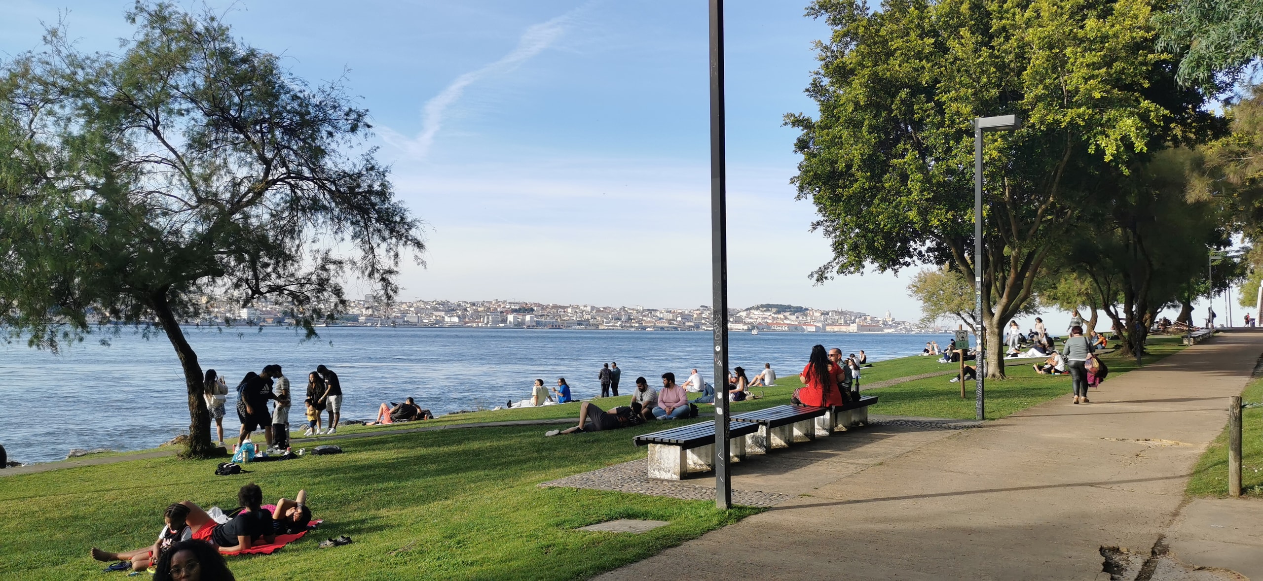 Young people sitting and lying on a grassy riverbank in Cacilhas, with a red picnic blanket, a paved path, and Lisbon across the Tagus
