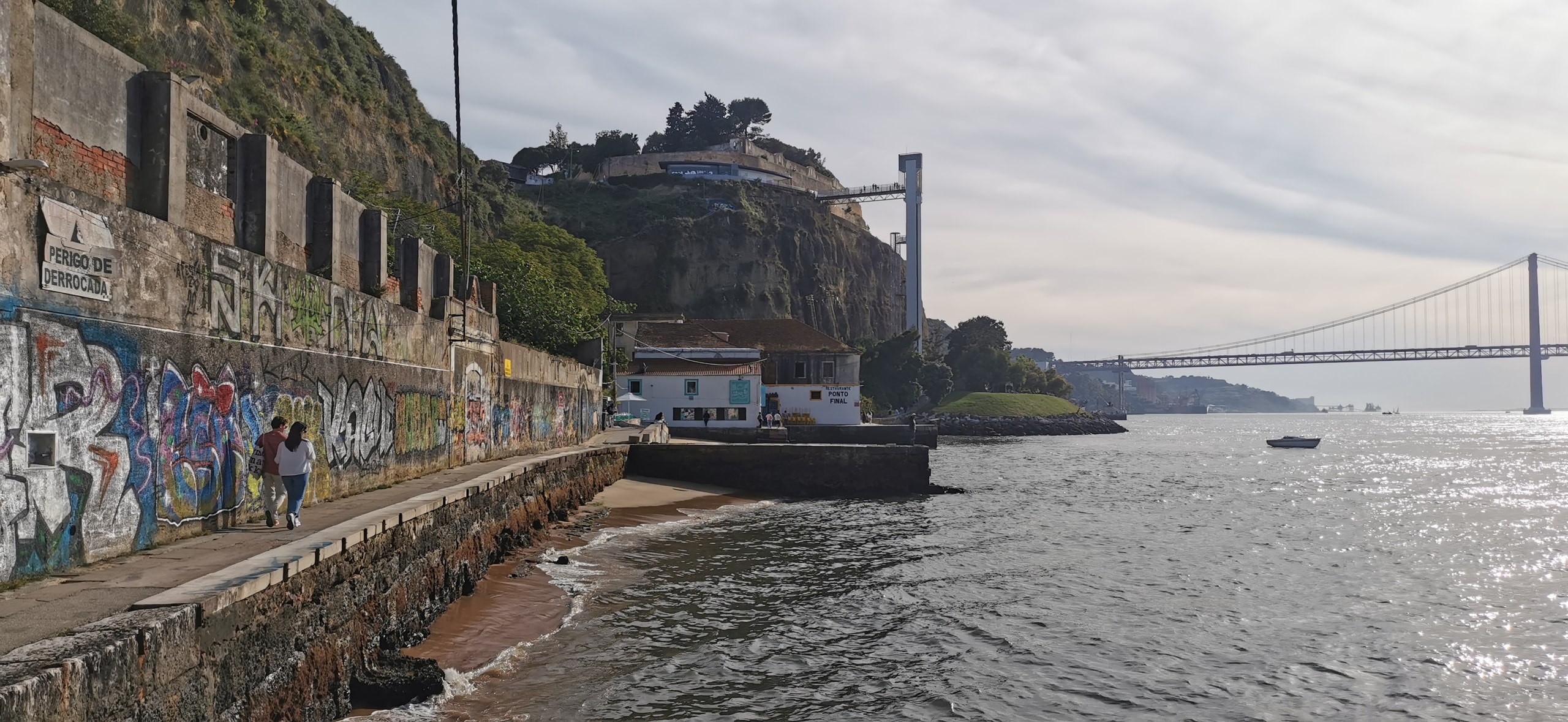 The river path at Cais do Ginjal in Cacilhas, with a graffiti wall, Restaurante Ponto Final ahead, and the 25 de Abril Bridge across the Tagus