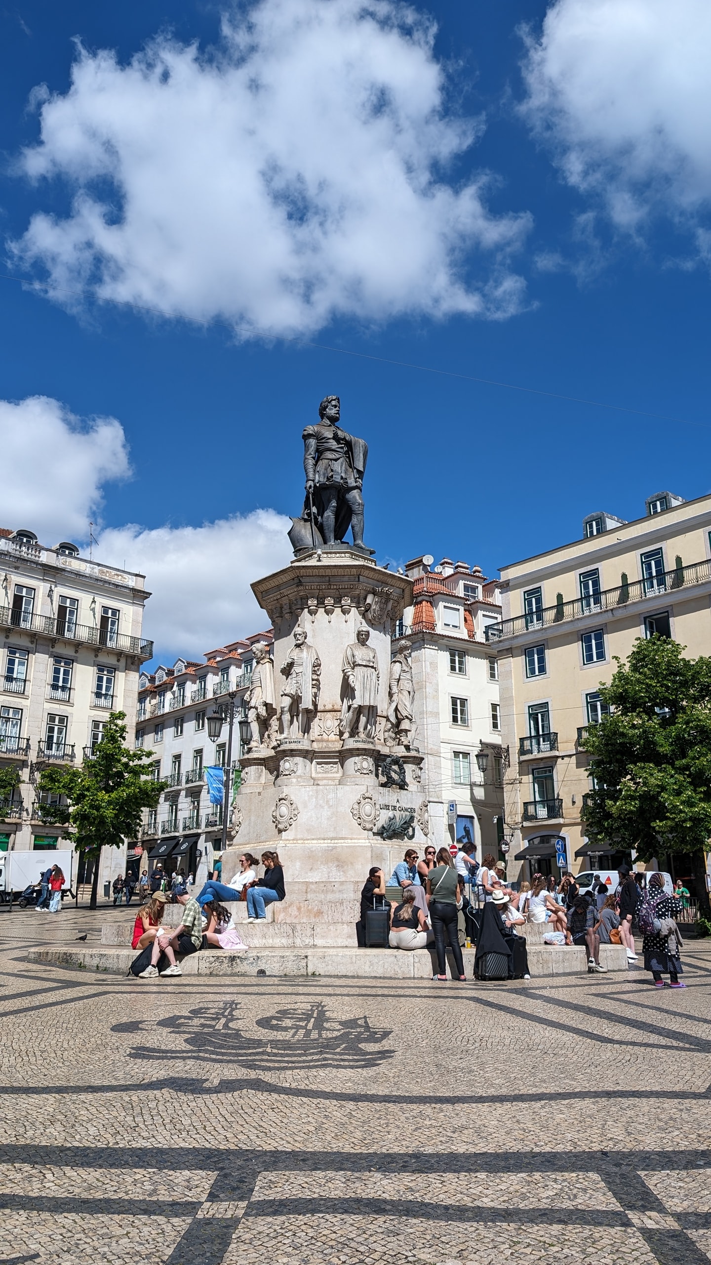 Bronze statue of Luís de Camões on a white pedestal in Chiado, with people sitting on the steps and a ship mosaic in the calçada