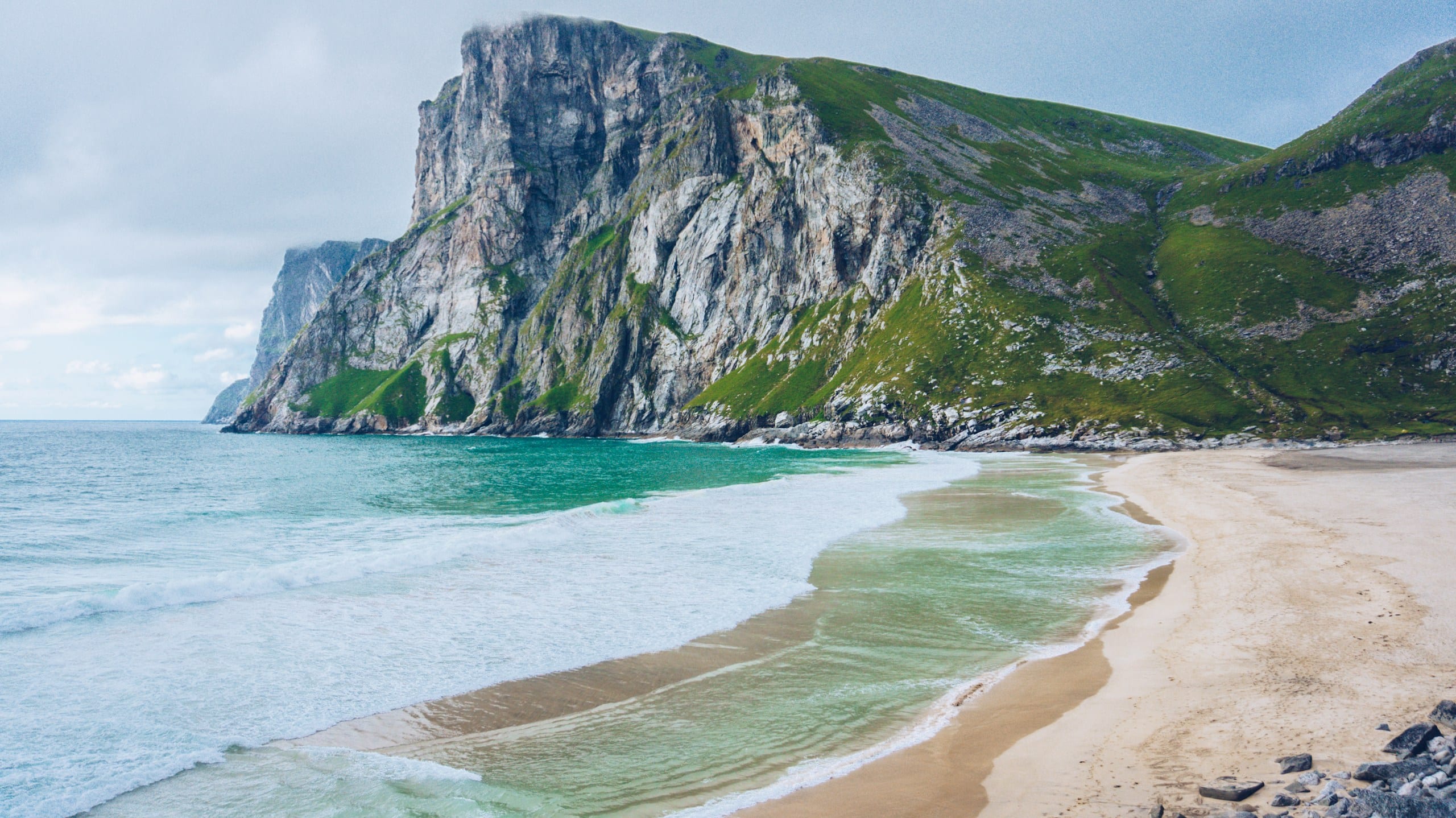 Pale sand and turquoise surf at Kvalvika Beach framed by steep green and grey cliffs in Lofoten