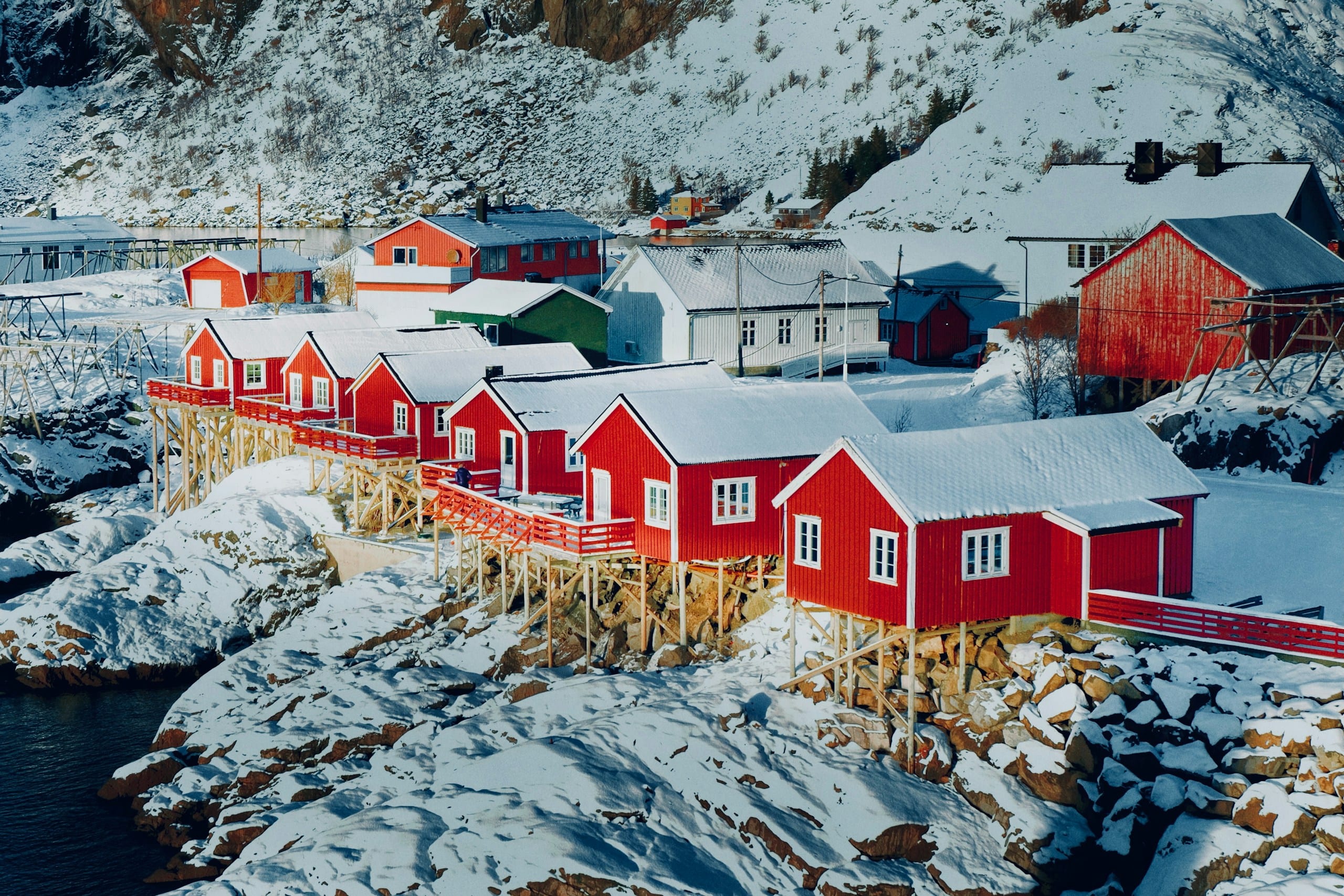 Red rorbu fishing cabins on stilts along a snowy rocky shore beneath a steep Lofoten cliff