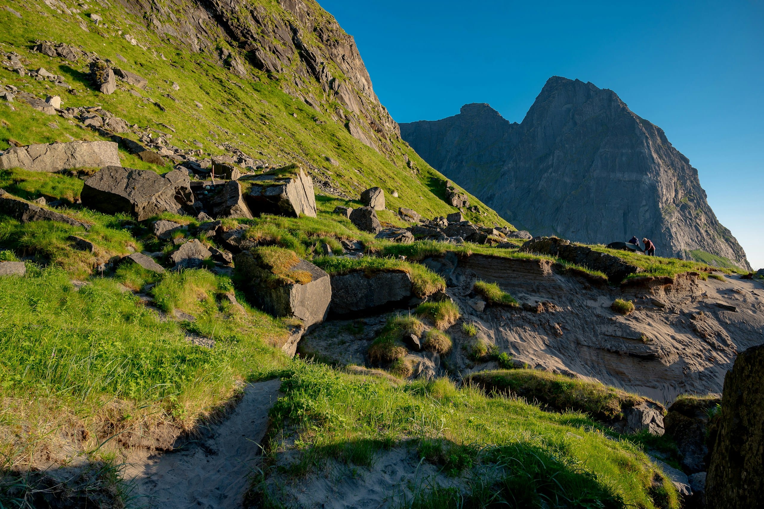 Grassy boulder slope and hiking path toward jagged Lofoten peaks under a clear blue sky