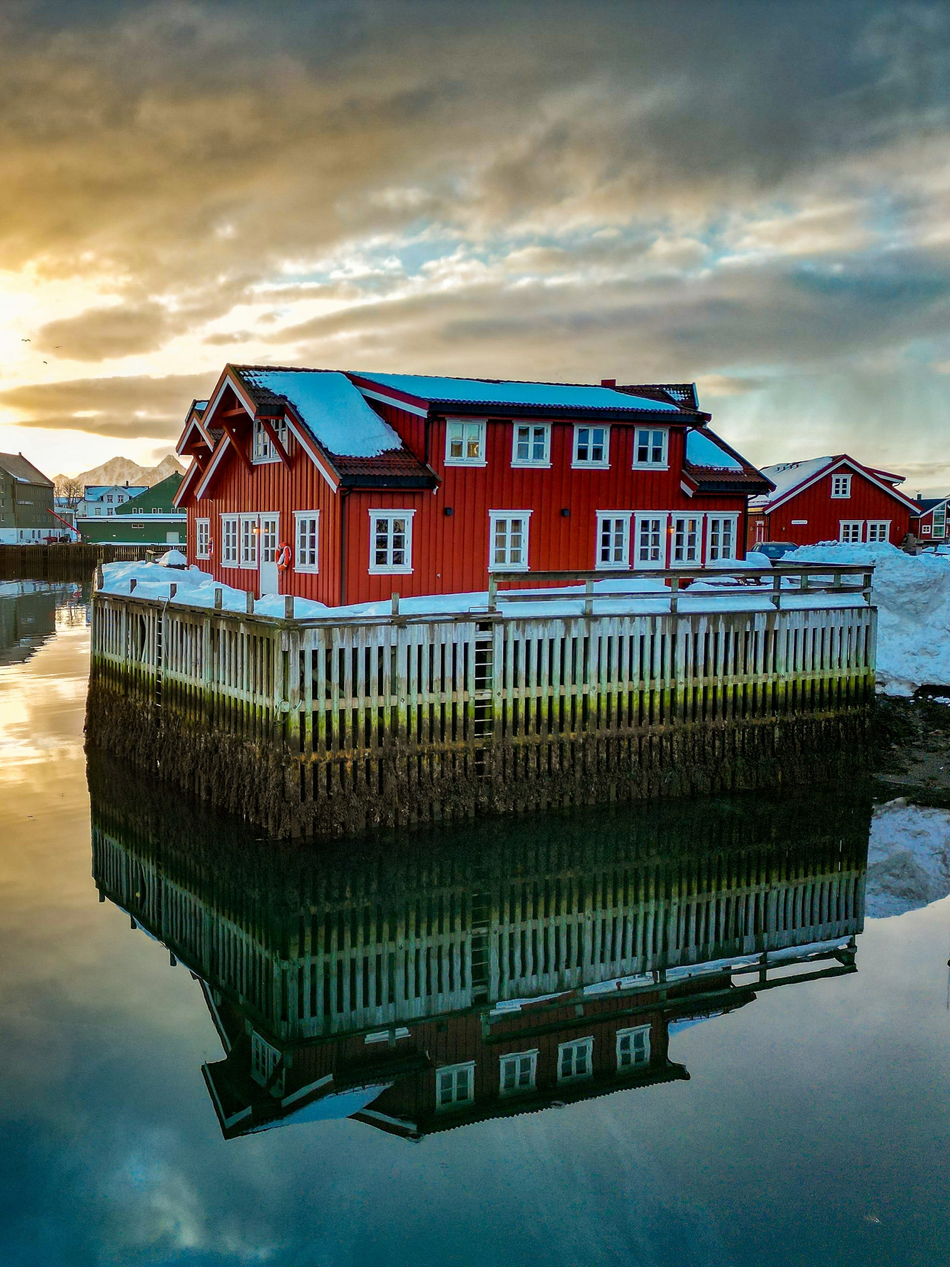 A red Lofoten rorbu on stilts reflected in calm harbour water with snow on the roof and mountains behind