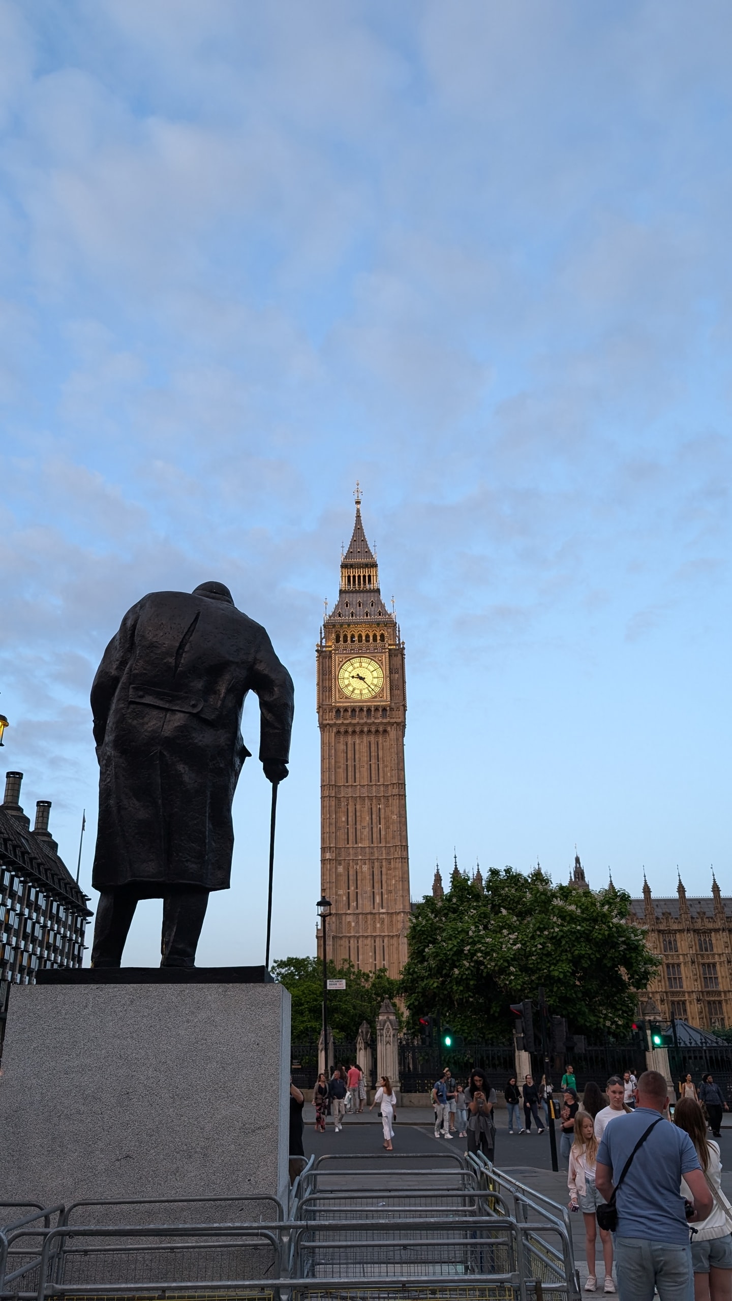 The dark bronze statue of Winston Churchill seen from behind in Parliament Square, leaning on a stick and facing Big Ben, whose clock face is lit gold against an evening sky