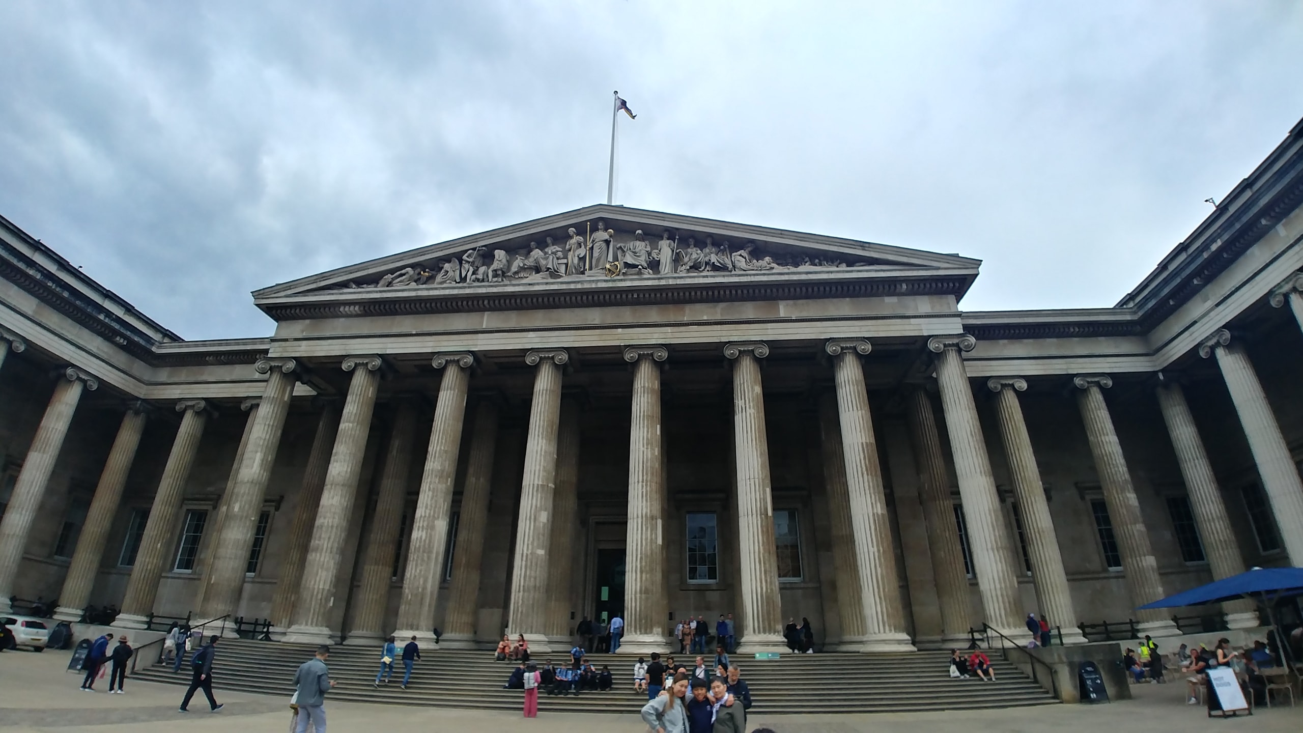 The British Museum's main entrance on Great Russell Street, a long row of tall stone columns under a carved triangular pediment, with visitors sitting on the wide front steps under a cloudy sky