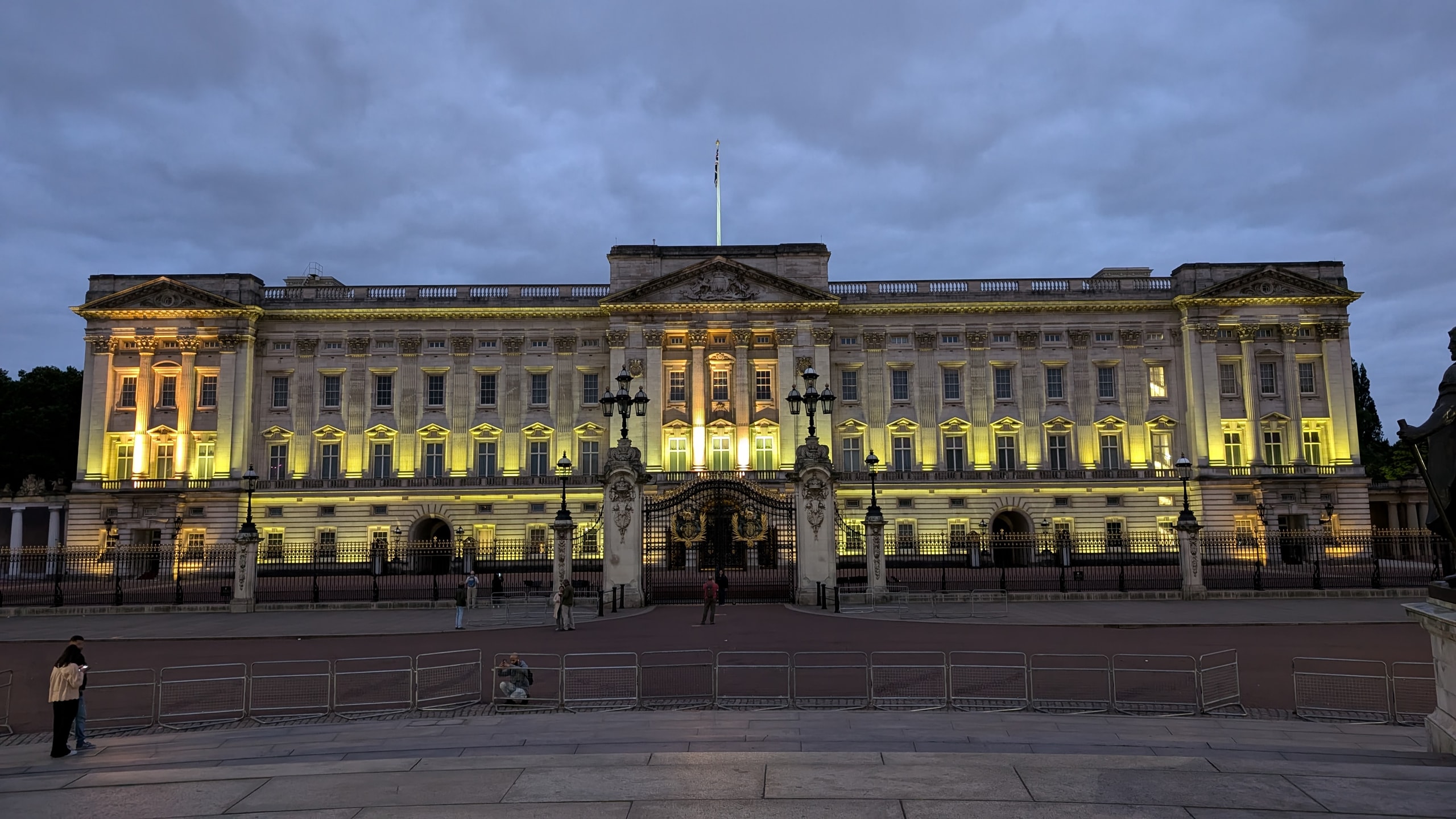 The front of Buckingham Palace floodlit in yellow at dusk under grey cloud, with its gold-trimmed iron gates, lamps and an almost empty forecourt behind metal crowd barriers