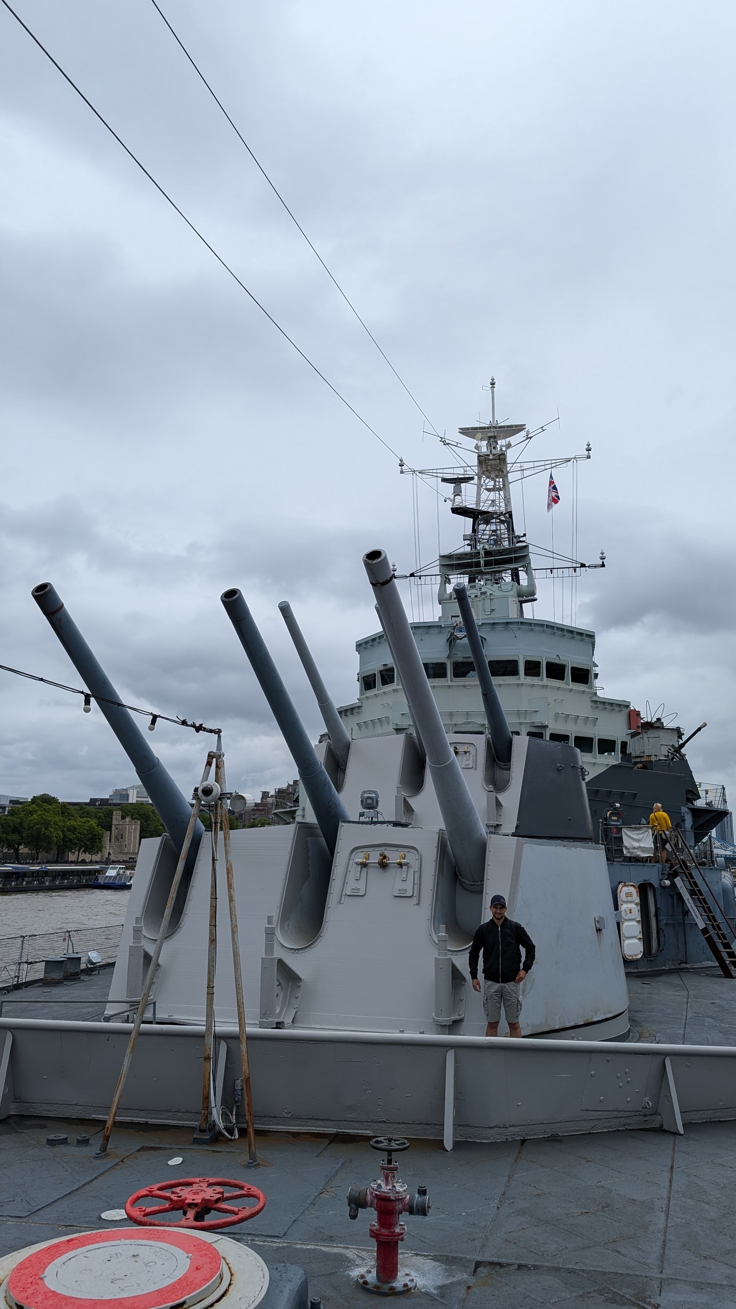 The grey forward gun turrets of HMS Belfast with their long barrels angled upwards, the ship's bridge and mast behind, a visitor standing beside a turret, and the White Tower of the Tower of London across the river on the left