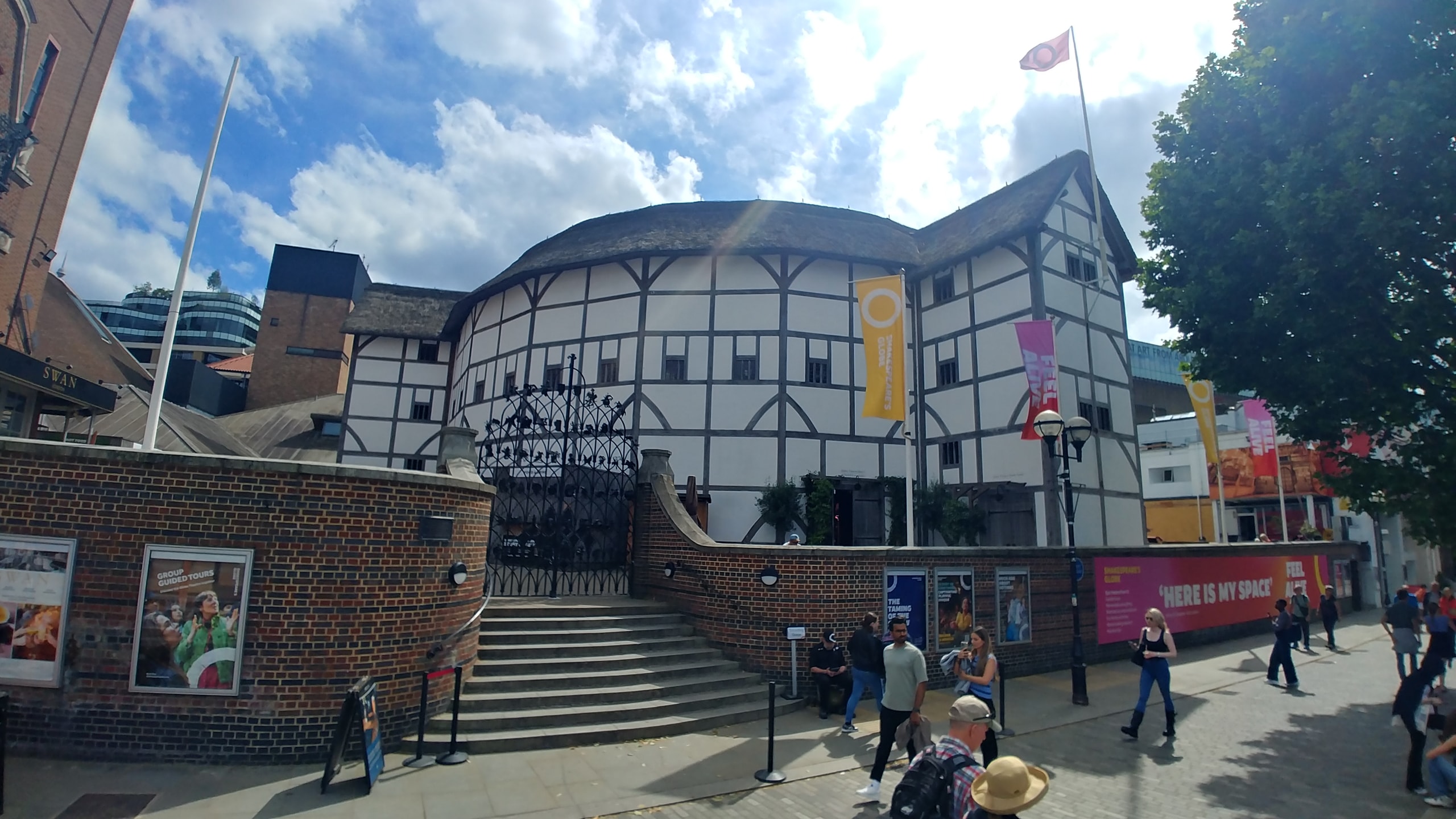 Shakespeare's Globe on the South Bank, a round white building with dark oak beams and a thatched roof behind a brick wall and iron gates, with play banners and people walking past on a sunny afternoon