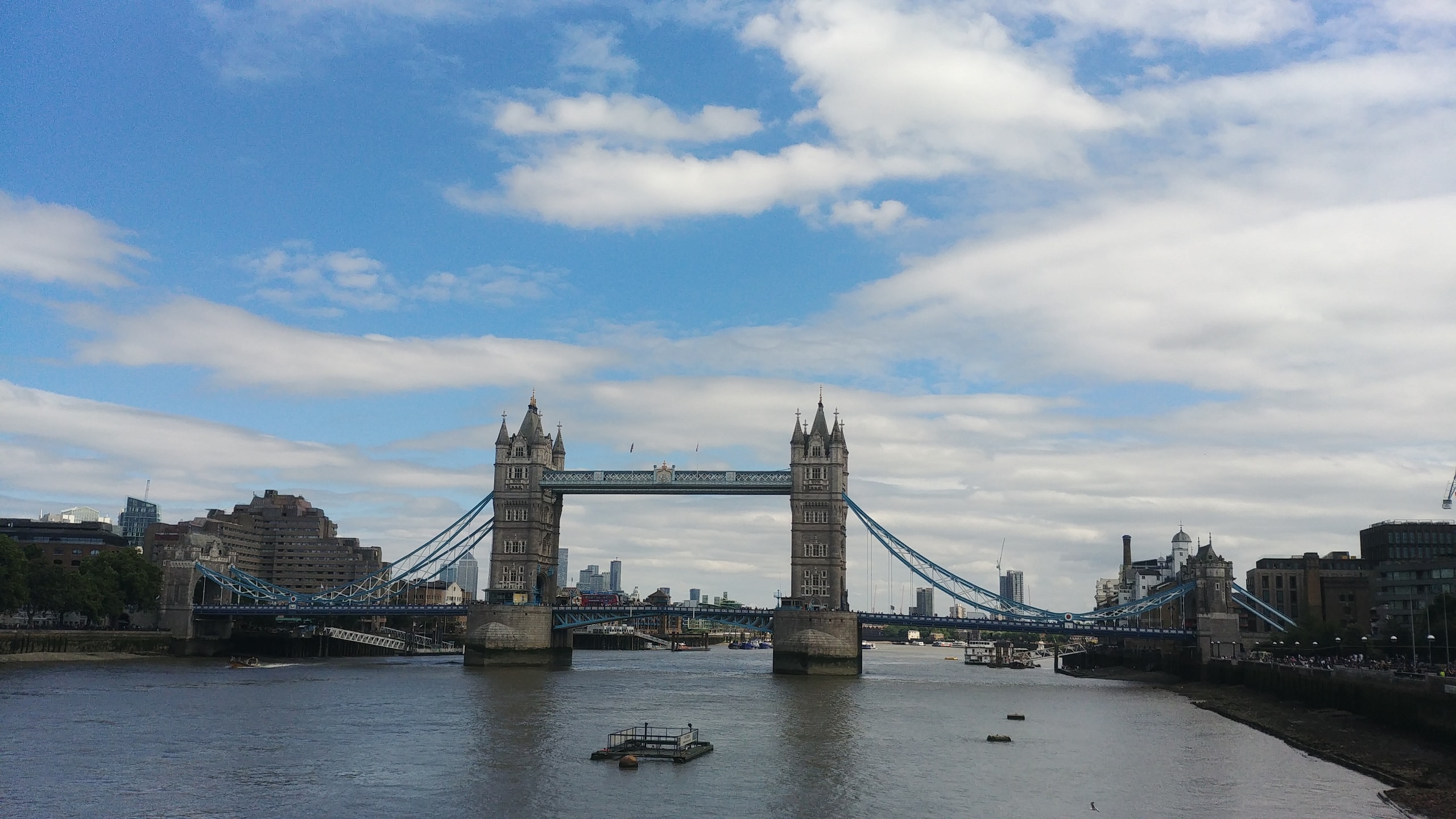 Tower Bridge across a wide stretch of the Thames under blue sky and white cloud, with its two stone towers, blue suspension chains and the Canary Wharf skyscrapers visible between the towers