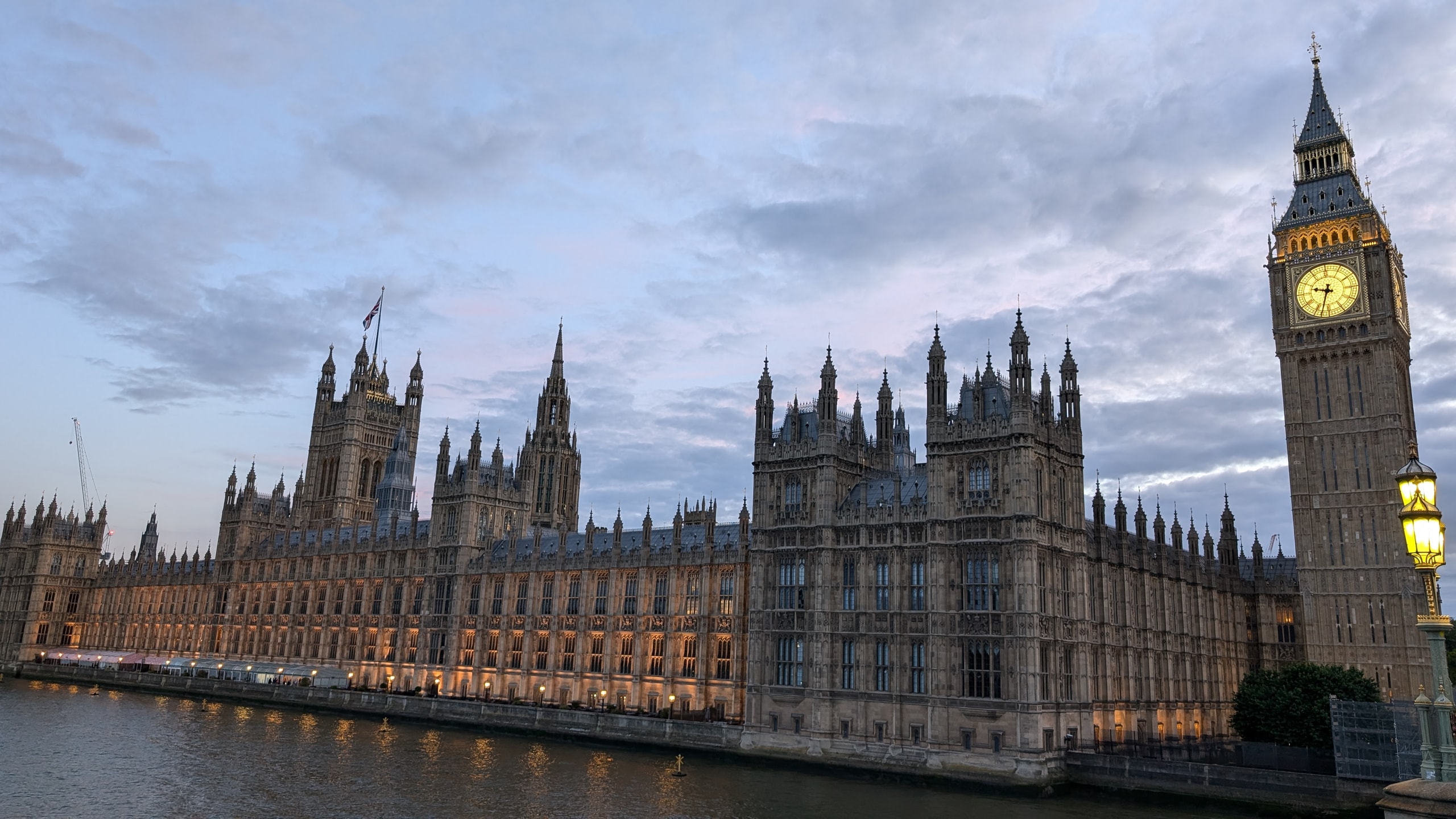 The river front of the Houses of Parliament at dusk, lit gold along the Thames, with the Victoria Tower flying a Union flag at the far end and Big Ben's lit clock face on the right