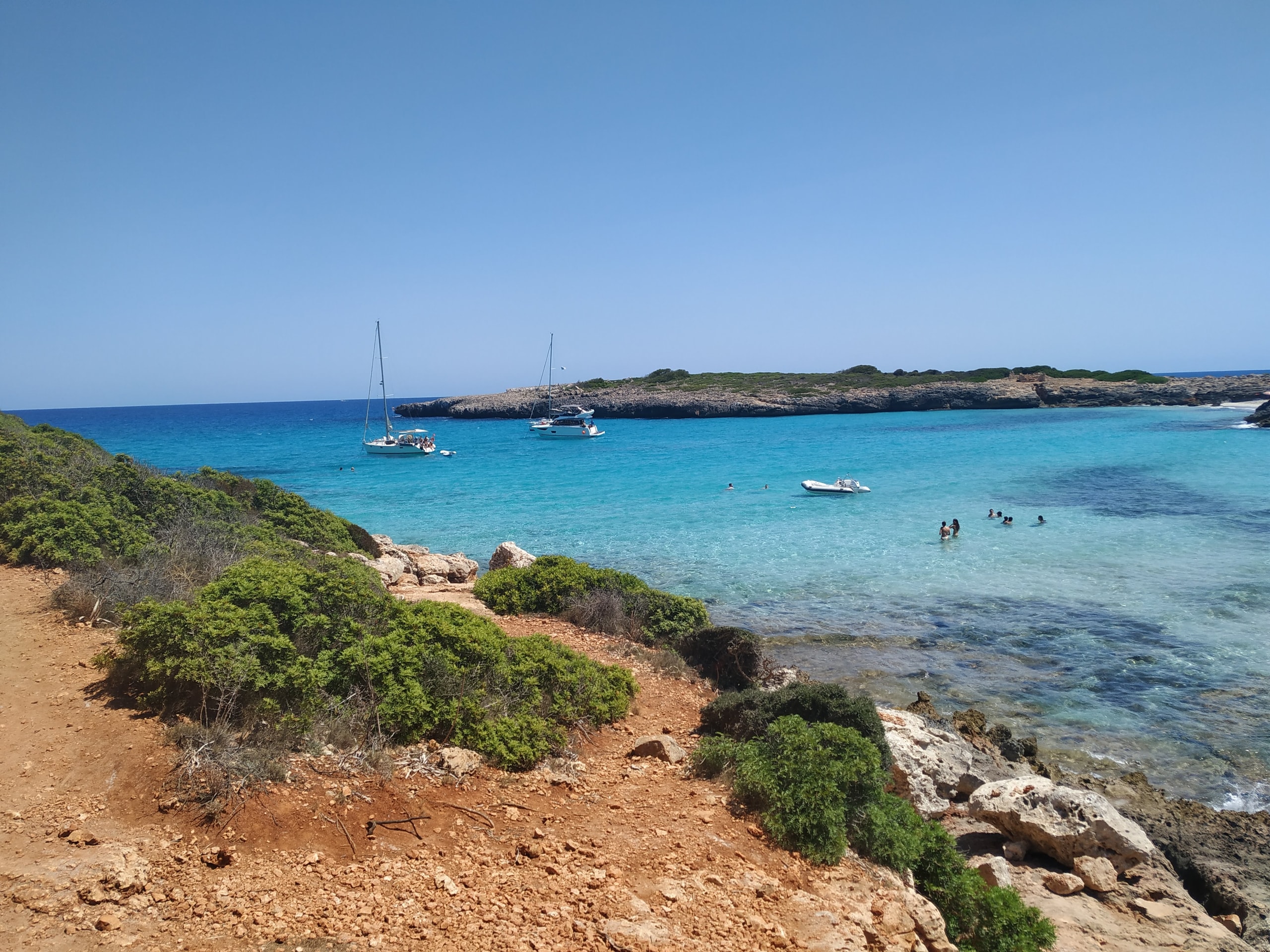 A rocky footpath beside clear water at Cala Varques with sailboats anchored across the bay