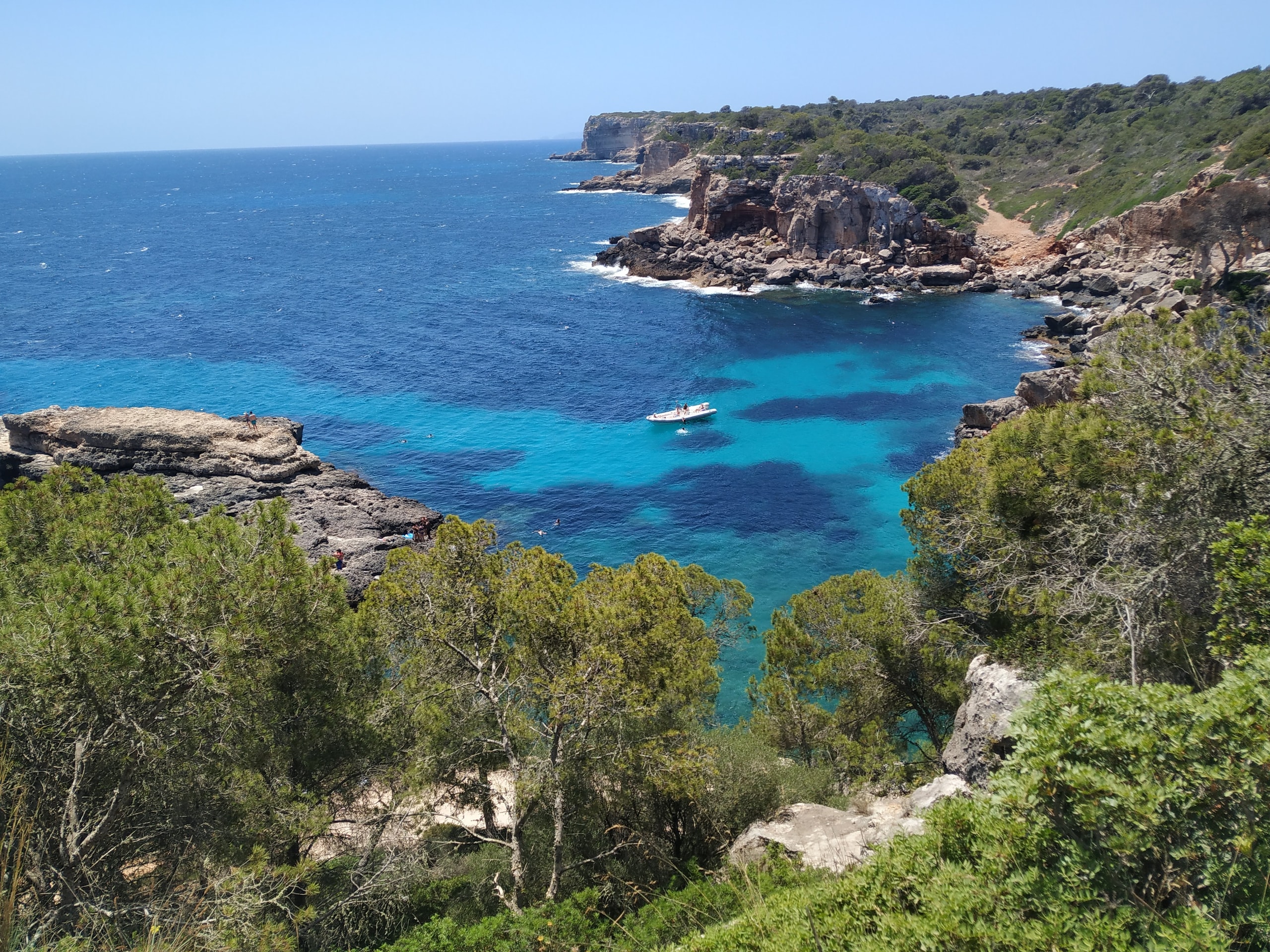 Clear turquoise water below pine-covered limestone cliffs at Cala Varques with a small boat anchored offshore