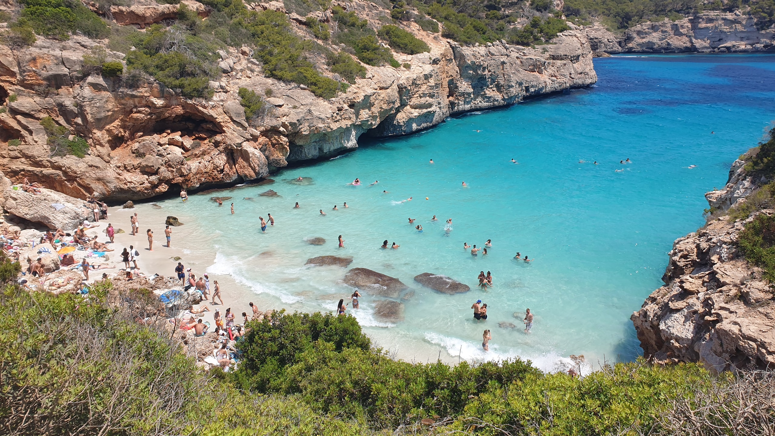 Caló des Moro crowded with swimmers and sunbathers between low limestone cliffs in July