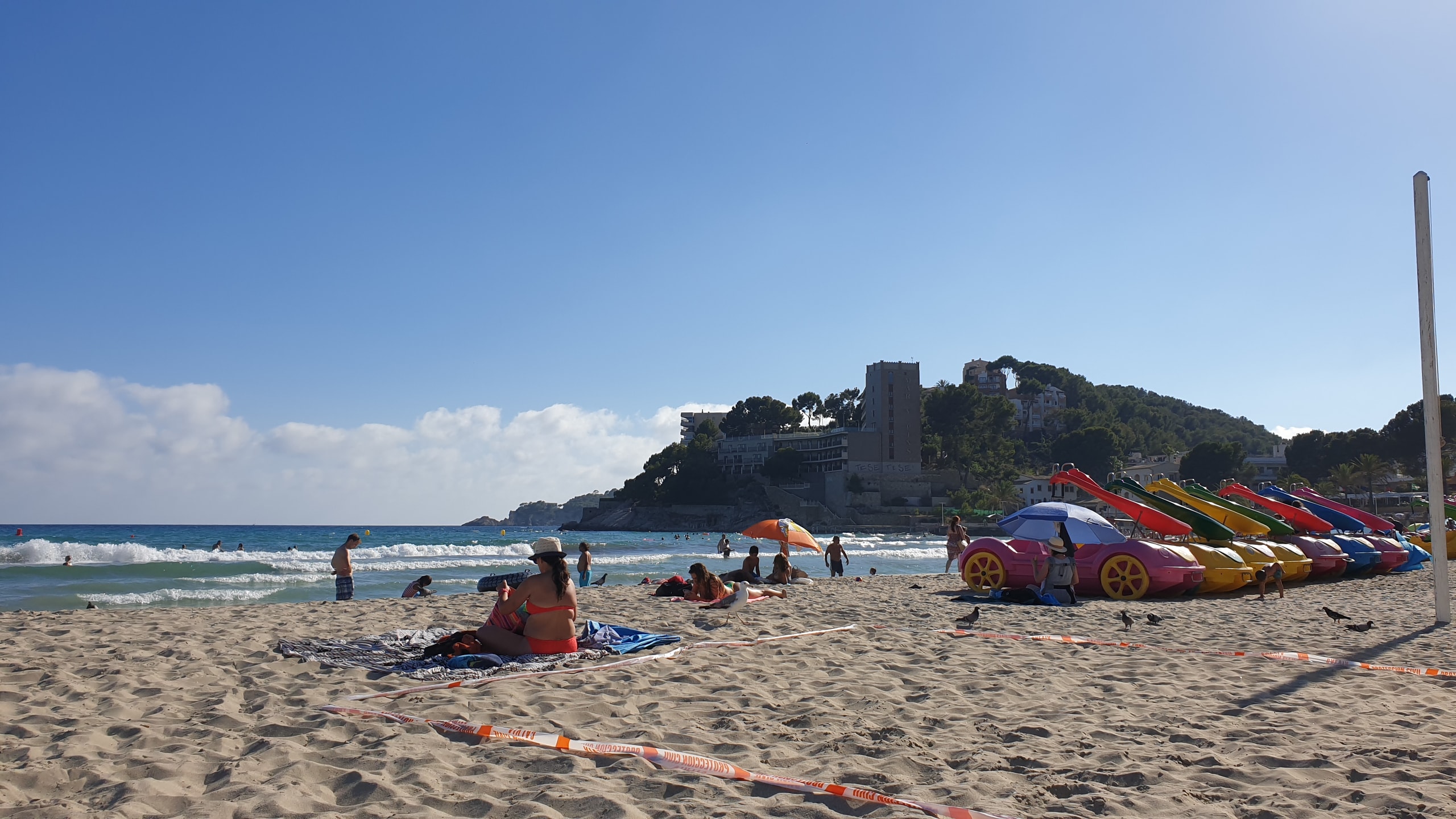 A busy sandy beach in Magaluf with swimmers, apartment blocks, and colourful pedal boats shaped like cars