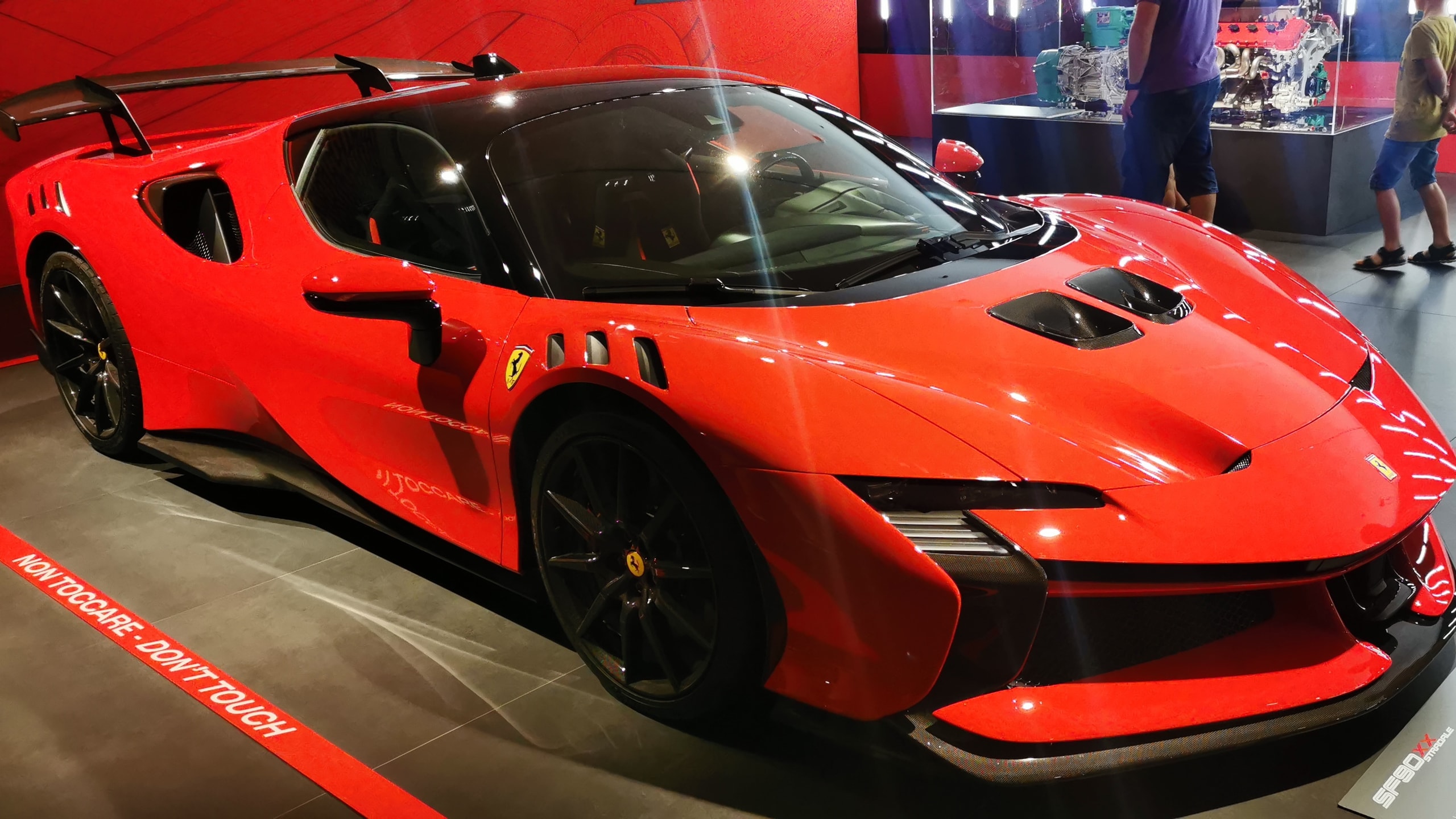 A red Ferrari SF90 XX Stradale with a large rear wing on display at the Museo Ferrari in Maranello, behind a red do-not-touch line on the floor